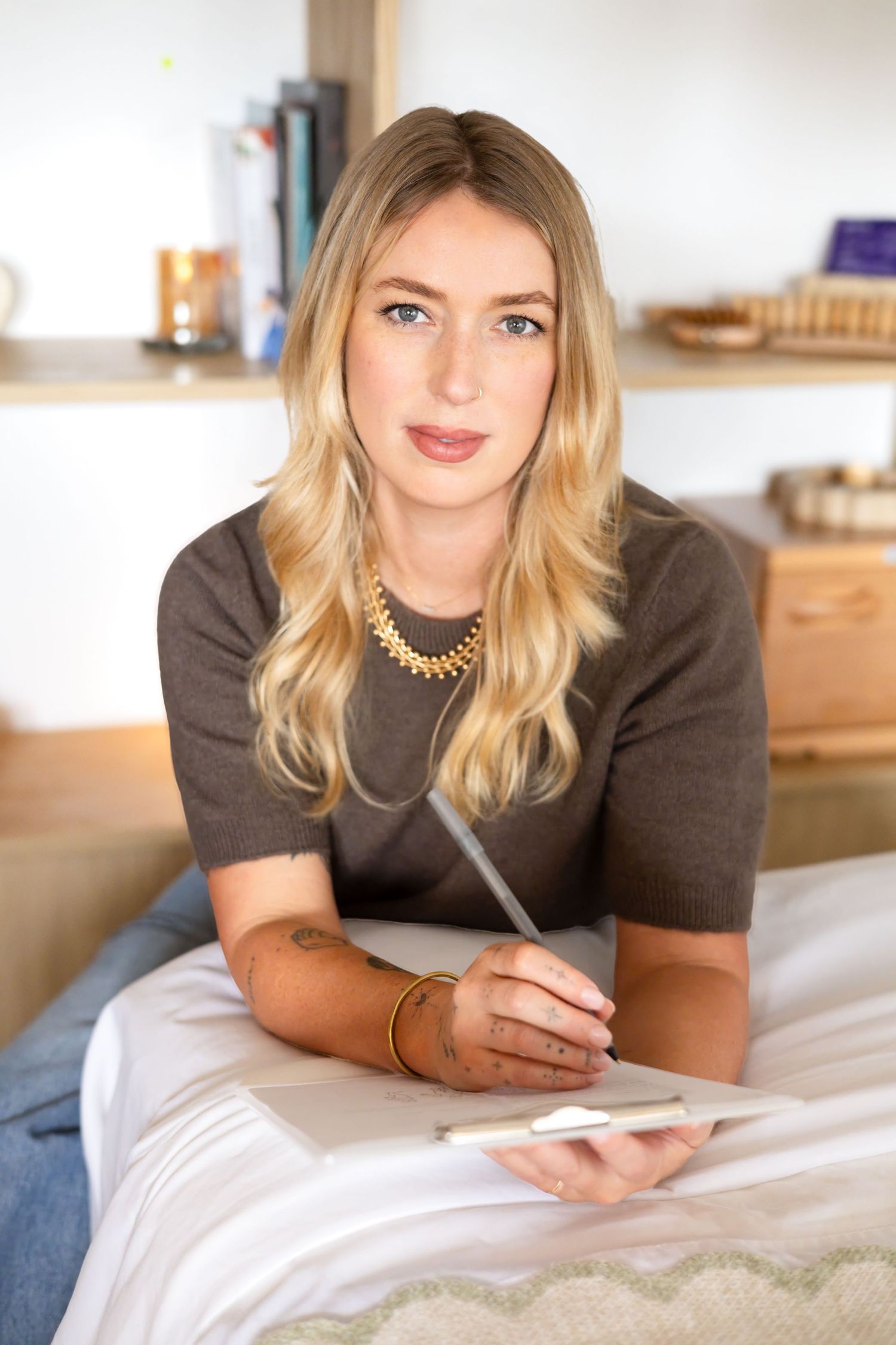 Smiling blonde woman with a gold necklace and tattooed forearm holds a pen and clipboard while seated on a bed in a bright wellness room.