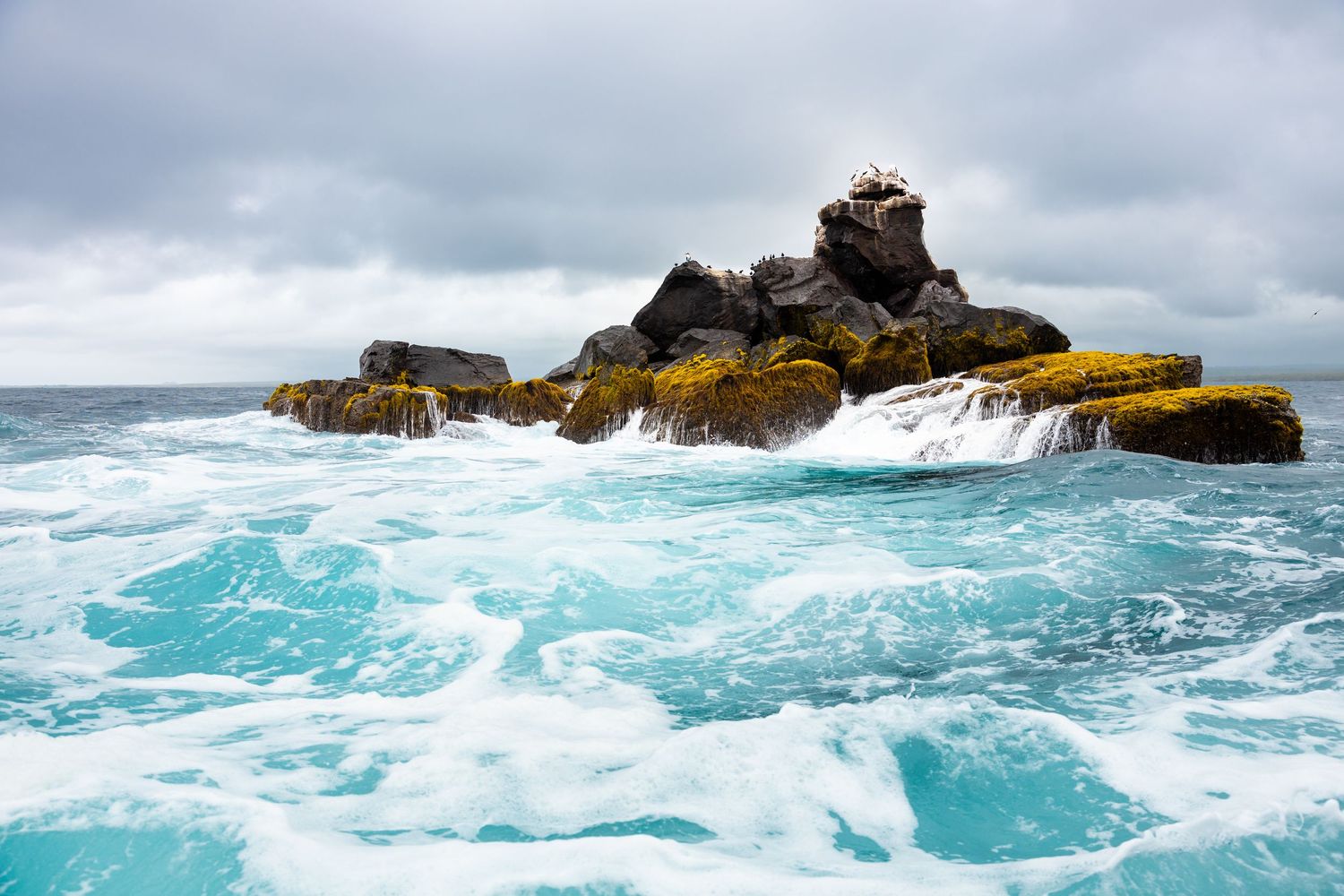 Rocky outcrop covered in moss and lichen rising from turquoise ocean water with white foamy waves under a cloudy sky.