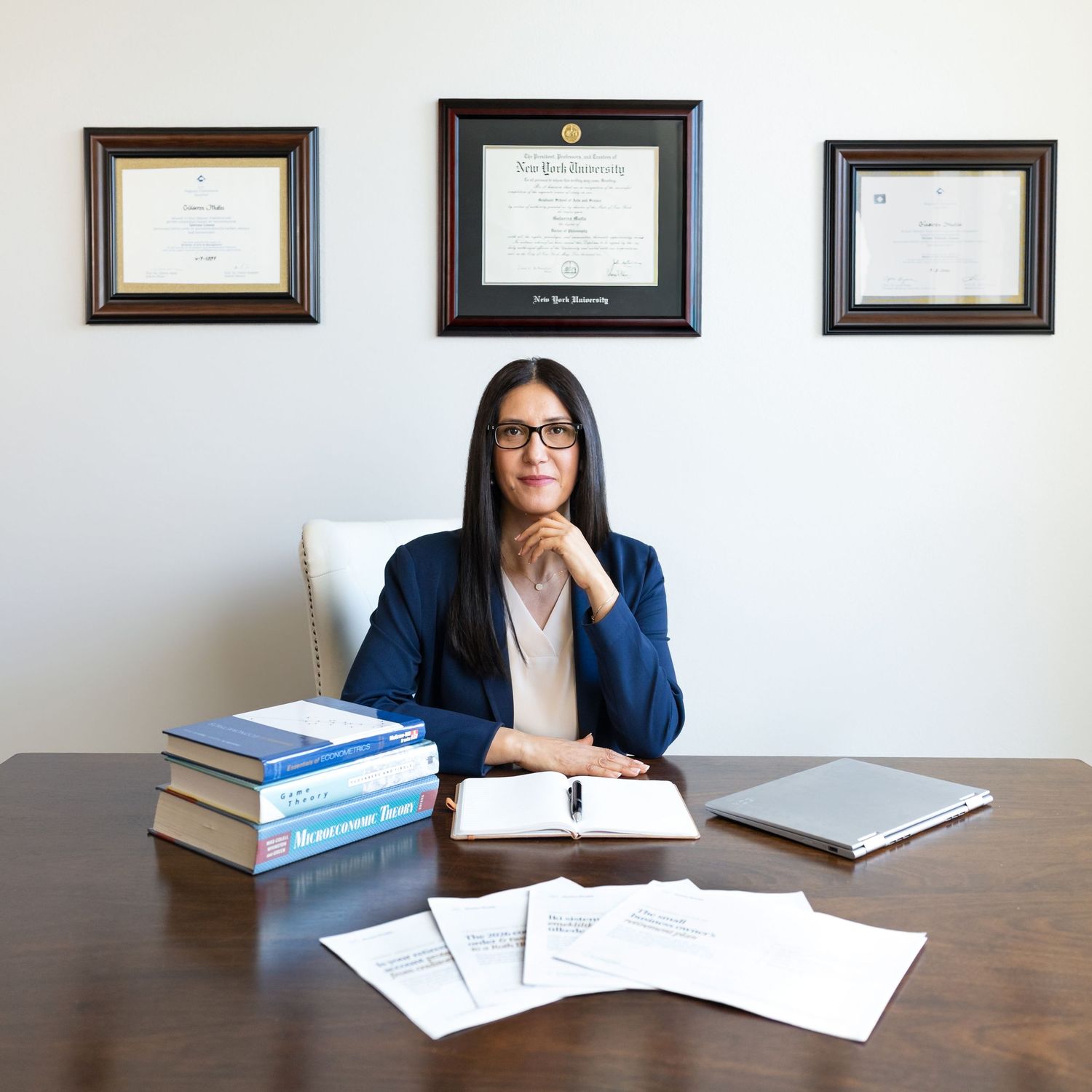 Woman in a navy blazer and glasses sits at a wooden desk with law books and papers beneath framed diplomas on the wall.