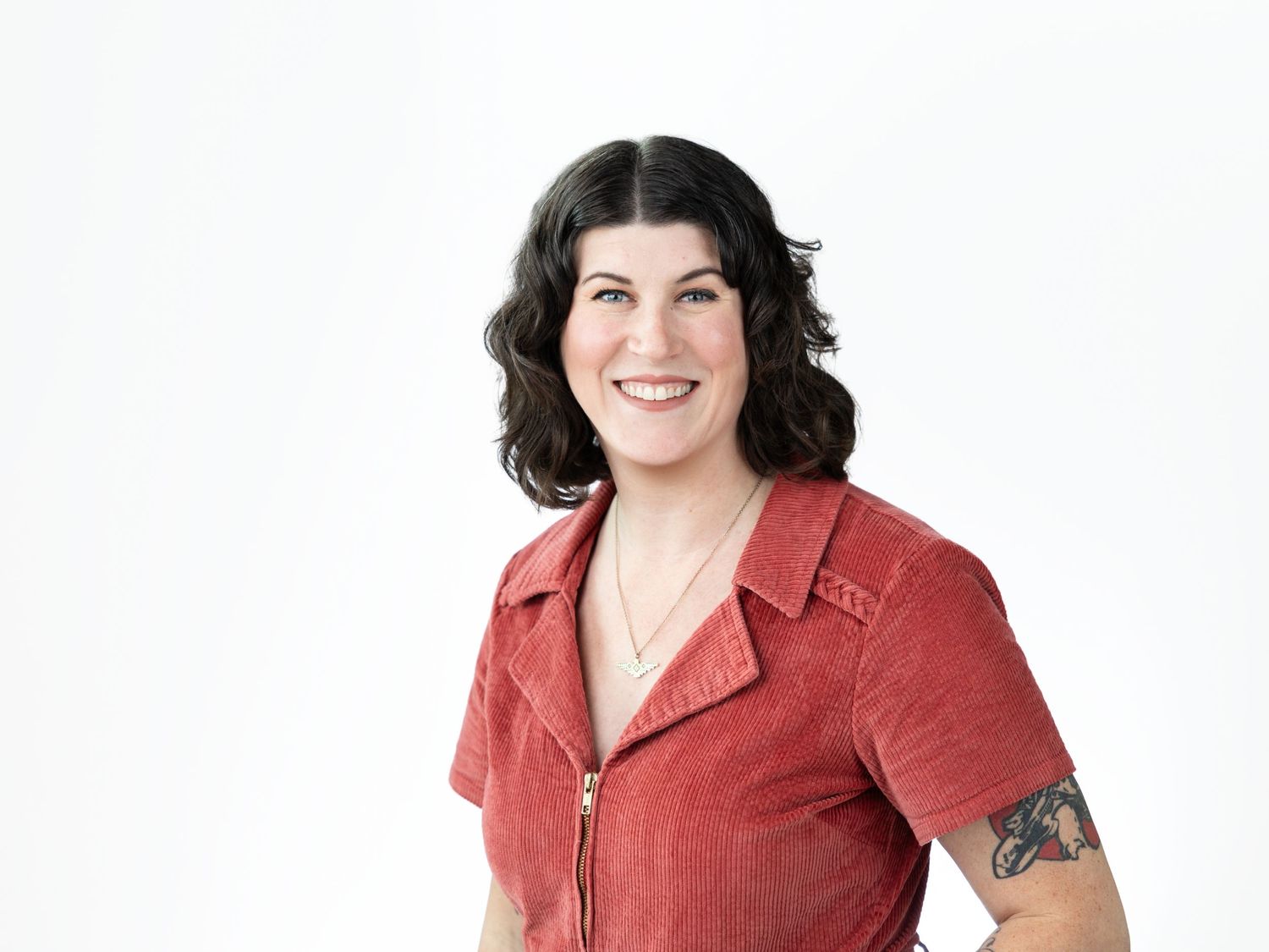 Smiling woman with short dark wavy hair and a forearm tattoo wearing a rust red corduroy zip-up top in a bright studio.