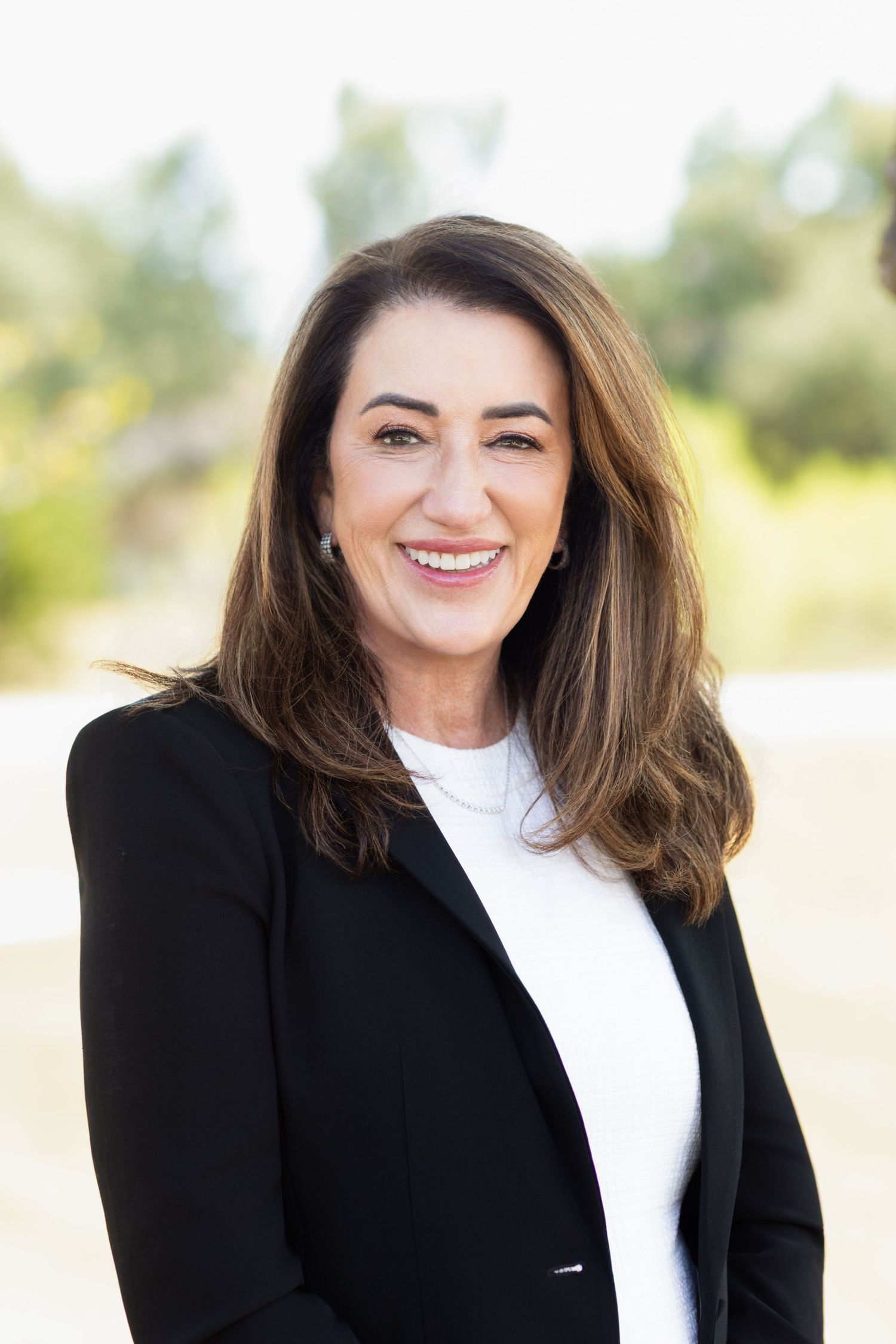 Smiling woman with shoulder-length brown hair wearing a black blazer and white top posing outdoors with a blurred natural background.