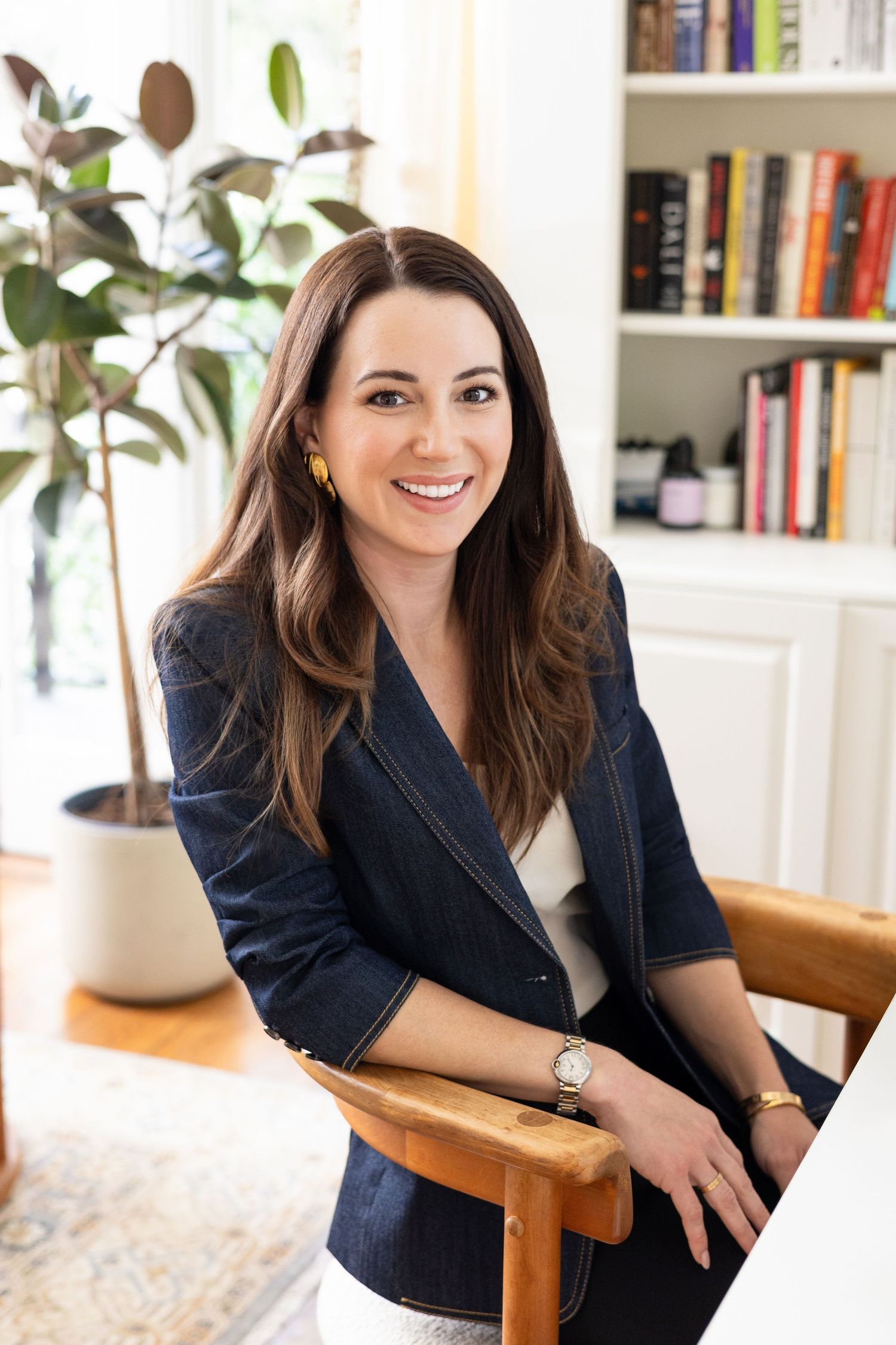A smiling woman in a navy denim blazer sits in a wooden chair in a bright home office with a plant and bookshelves.