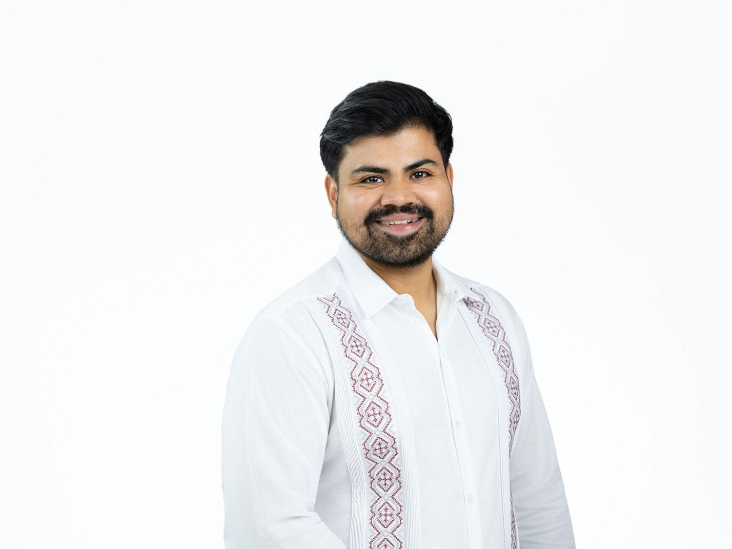 Smiling bearded man with dark hair wears a white shirt with purple embroidered detail in a professional portrait on a white background.