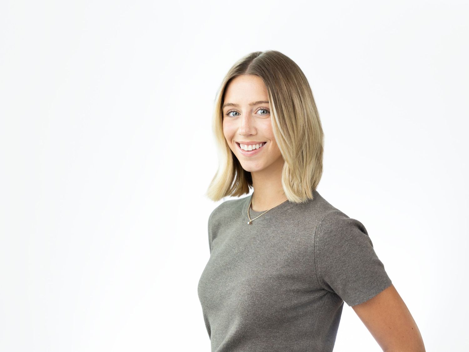 Smiling woman with a blonde bob haircut wearing a gray short-sleeve top and a thin necklace against a white background.
