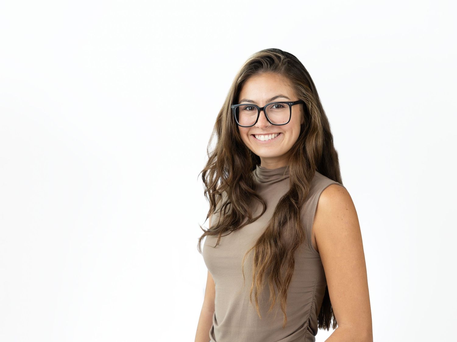 Smiling woman with long wavy brown hair and black-framed glasses wearing a taupe sleeveless turtleneck top against a white background.
