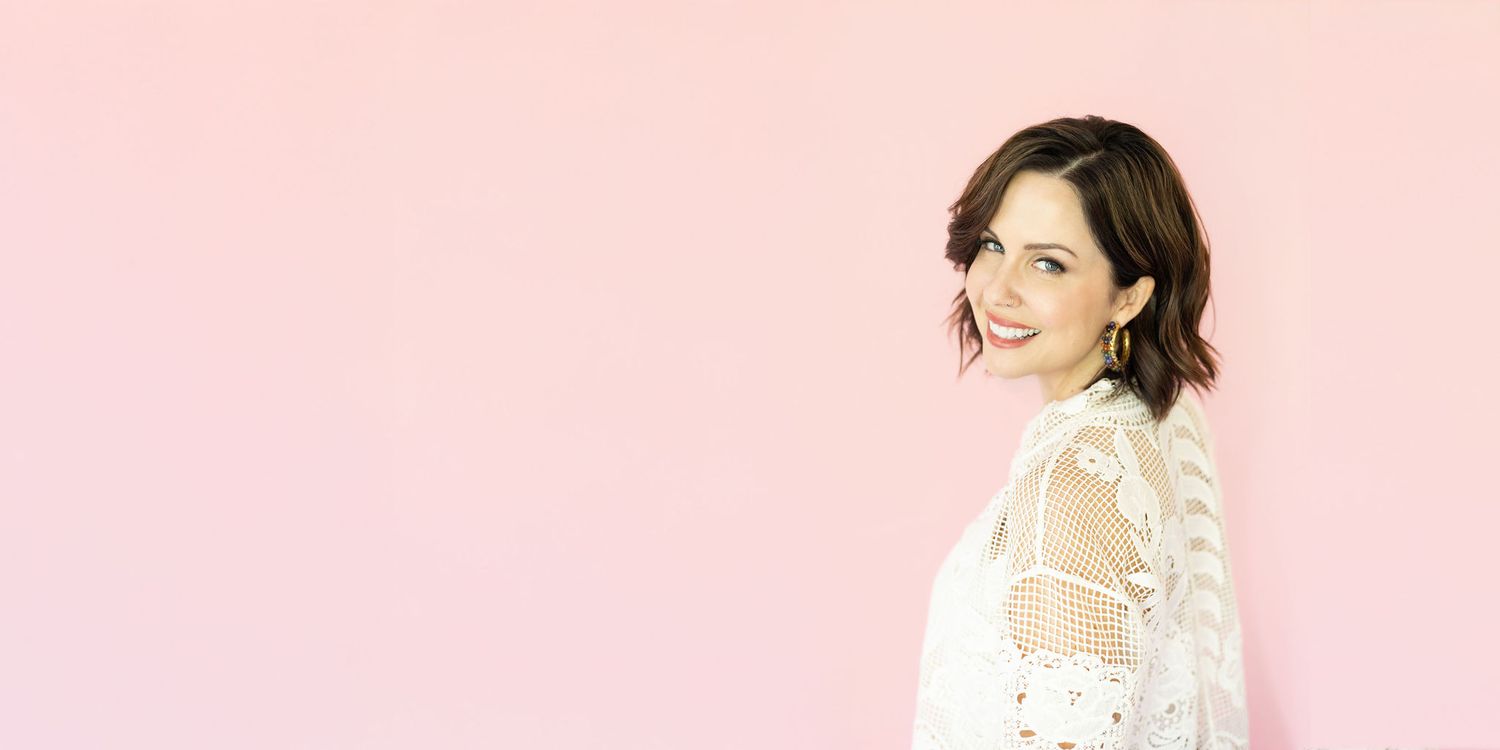 Smiling woman with short dark wavy hair wearing a white lace embroidered top, posed in profile against a soft pink background.