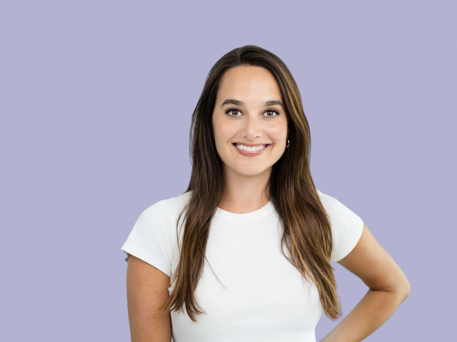 Smiling woman with long brown hair in a white t-shirt stands with a hand on her hip against a lavender background.