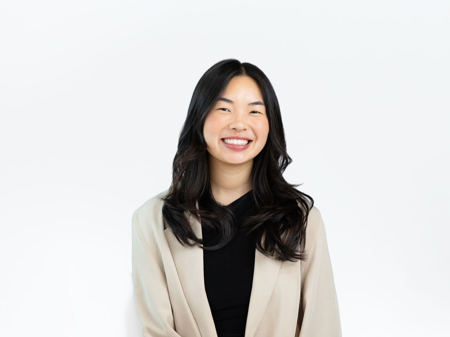 Smiling woman with long wavy black hair wears a cream blazer over a black top in a professional portrait on a white background.