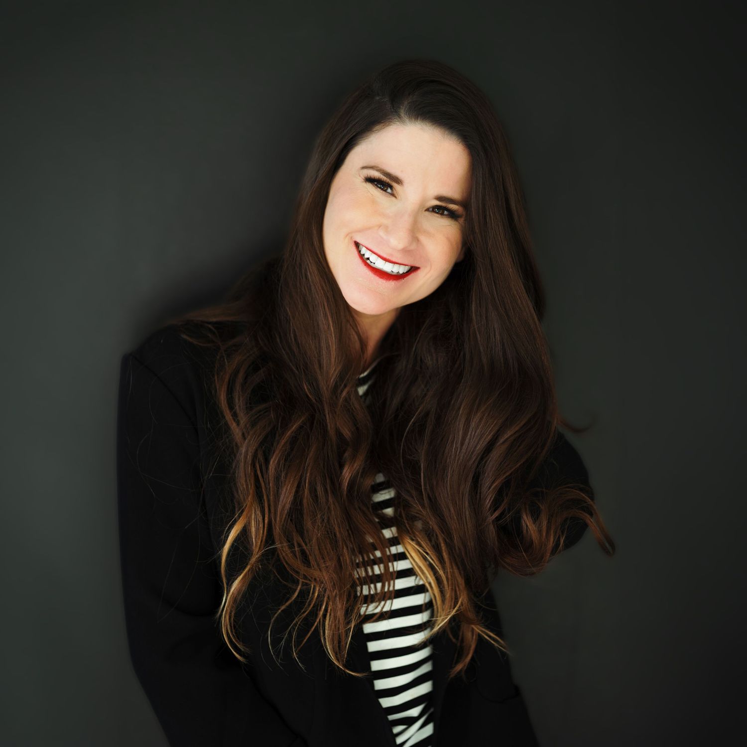 Smiling woman with long wavy brunette hair and red lipstick wears a black jacket over a striped top against a dark backdrop.