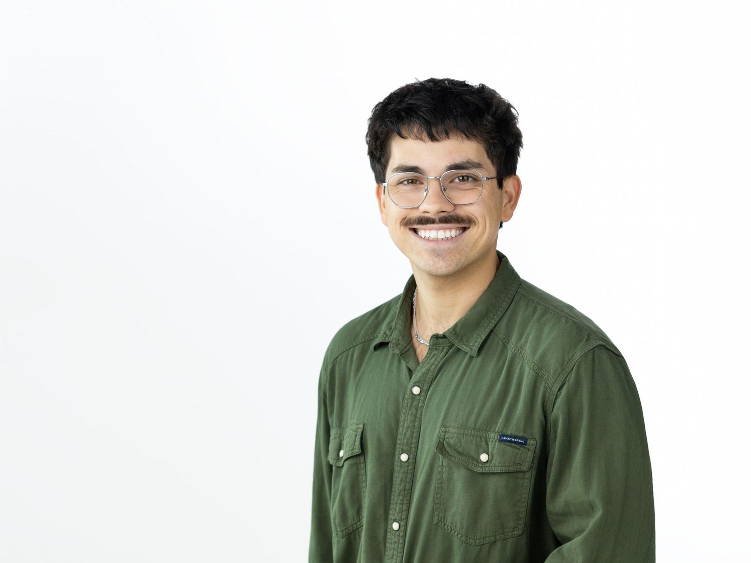 Young man with curly dark hair, glasses and a mustache smiling in an olive green button-up shirt.