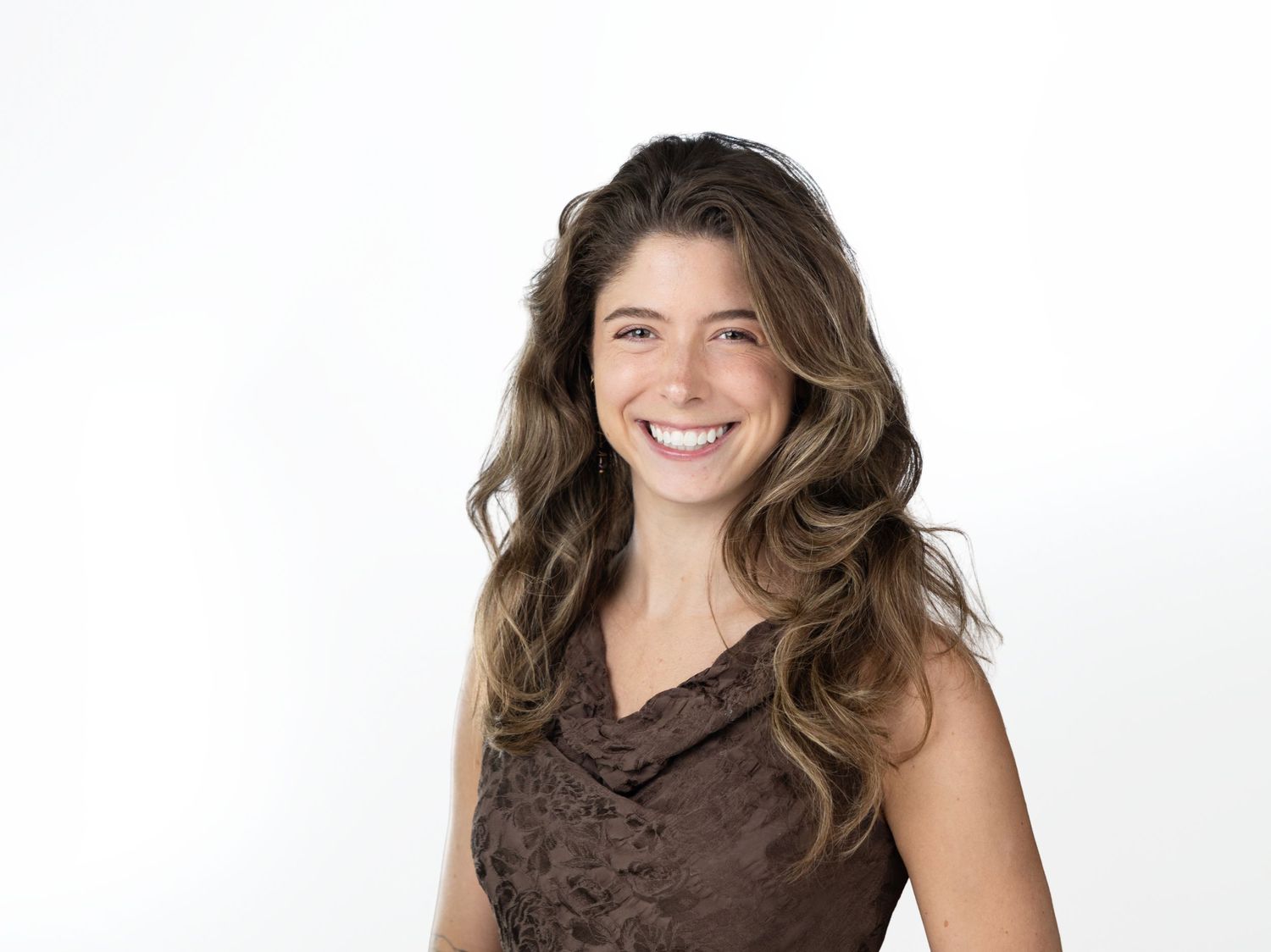 Smiling woman with long wavy brown hair wearing a brown textured sleeveless top in a bright, clean studio setting.