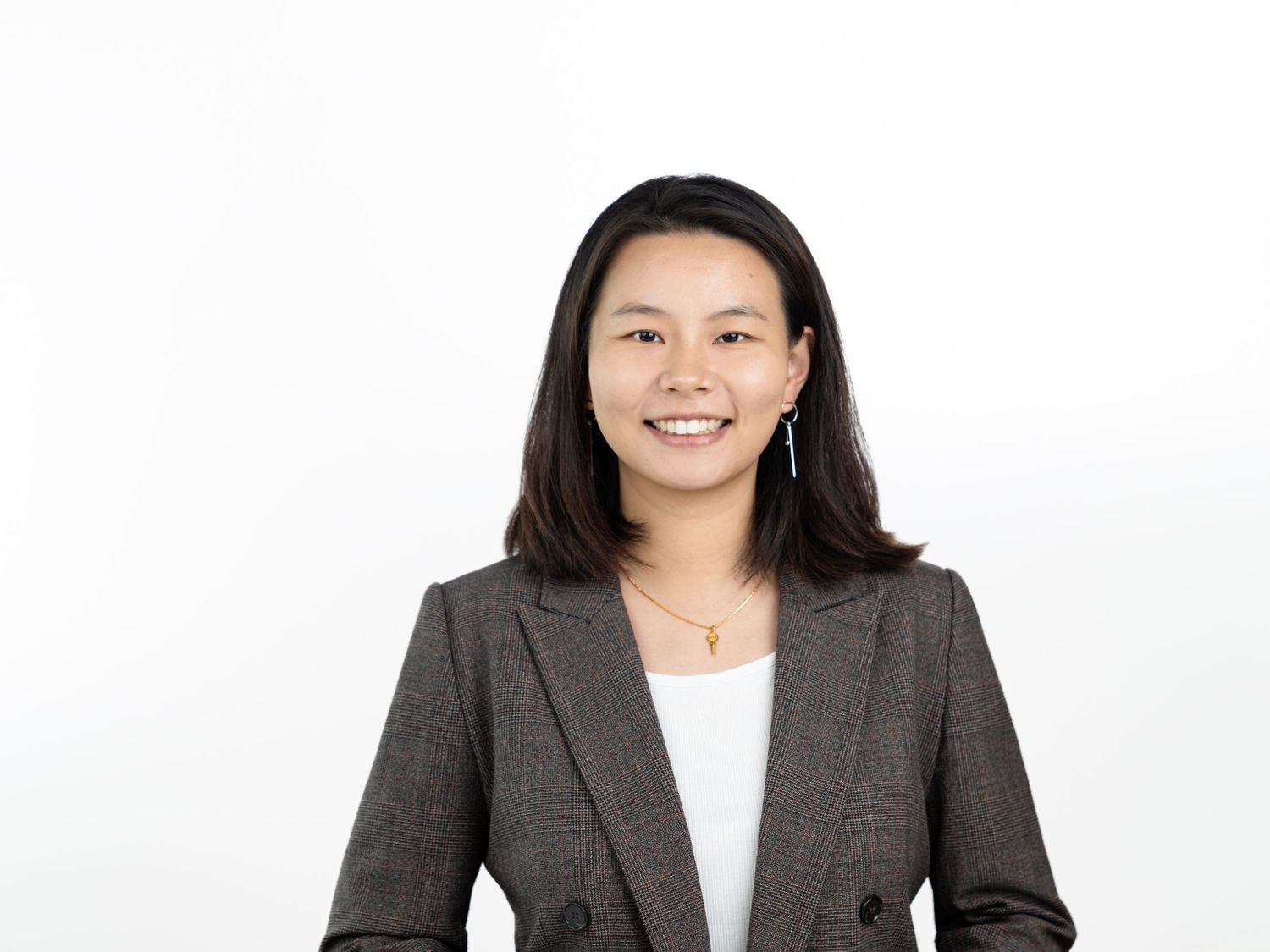 Smiling woman with straight dark hair wearing a gray tweed blazer over a white top and a gold necklace in a bright studio.