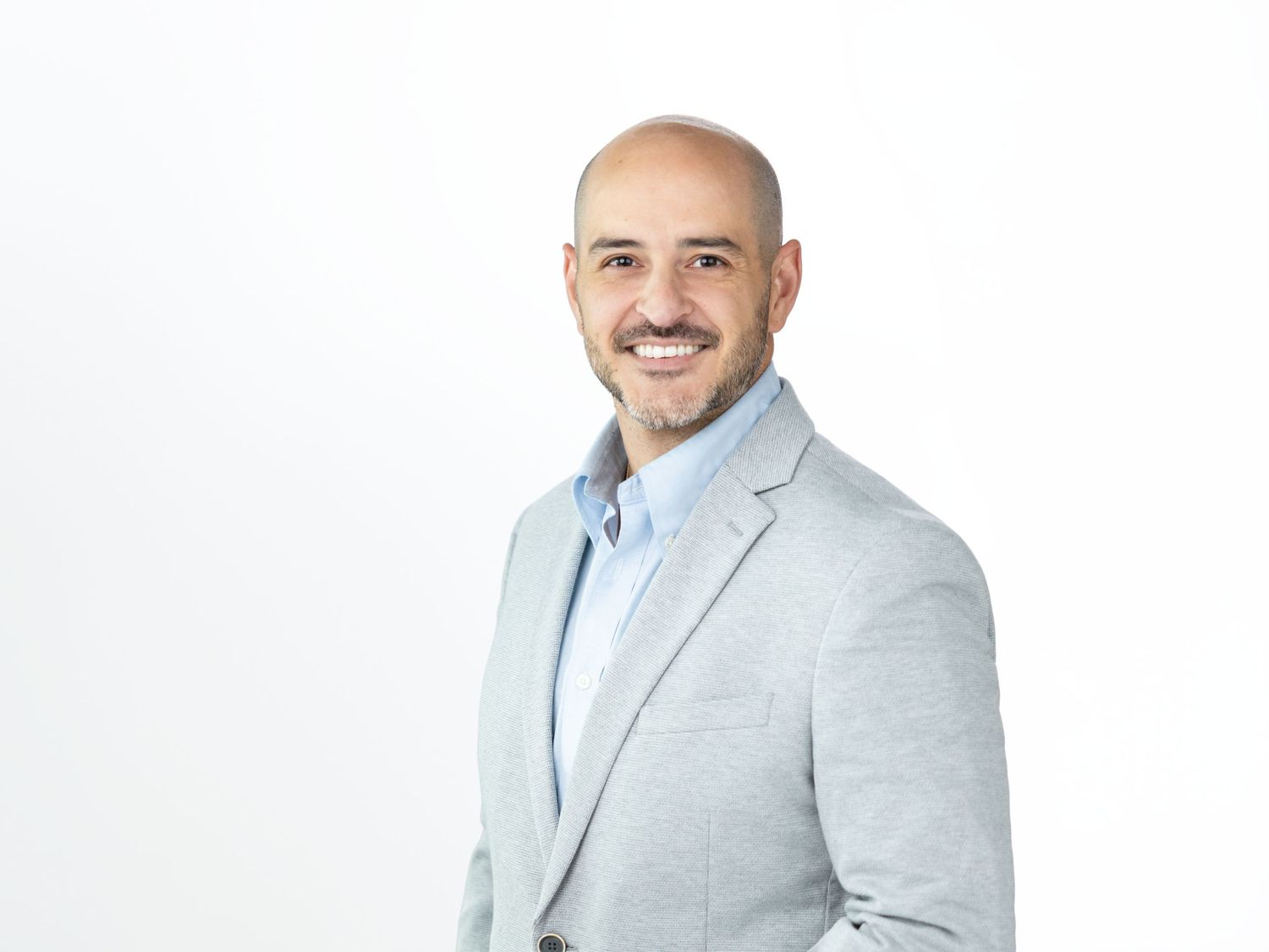 Smiling bald man with a trimmed beard wearing a light gray blazer over a light blue shirt against a white background.