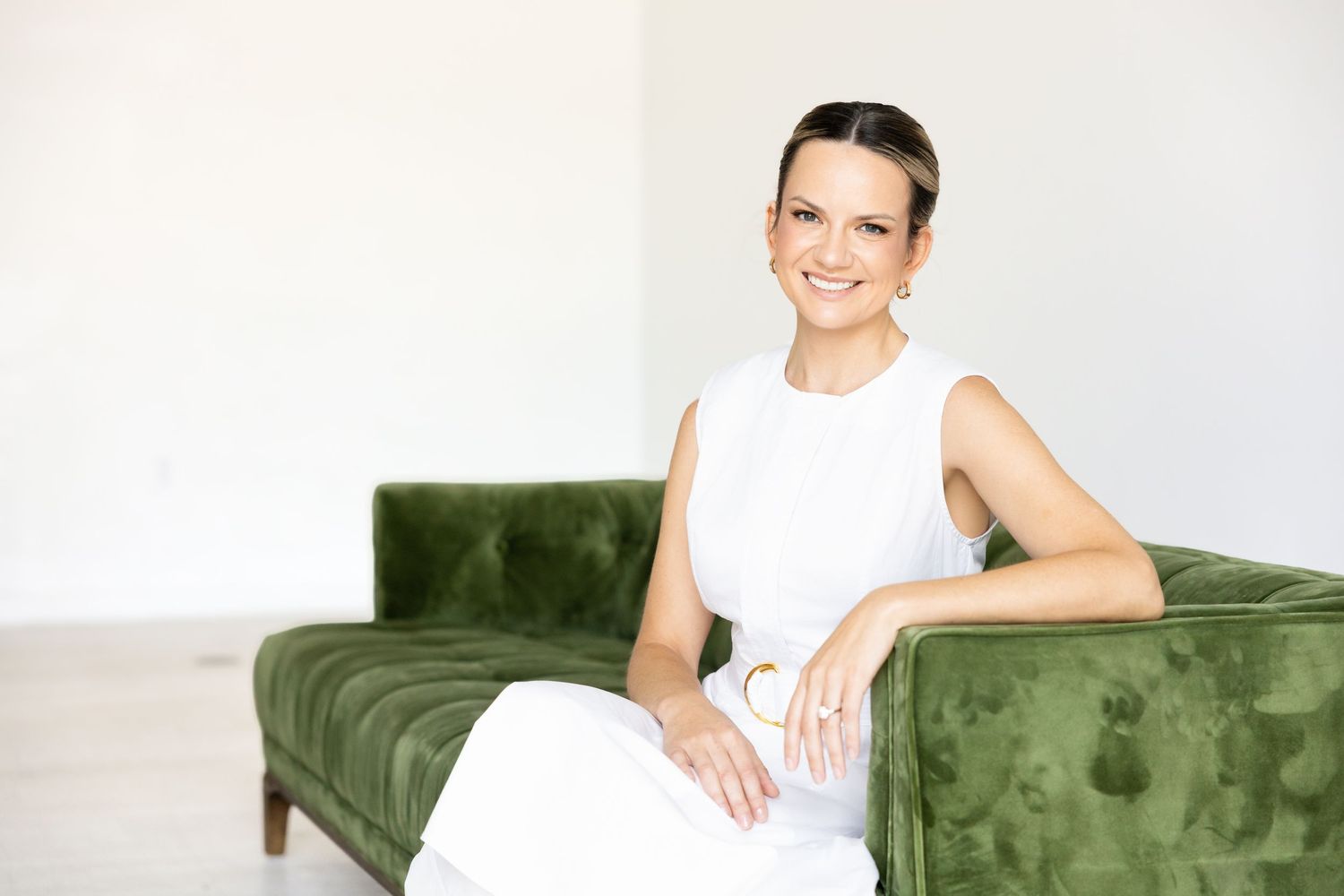 Smiling woman in a white dress sits gracefully on a green velvet sofa in a bright, minimalist room.