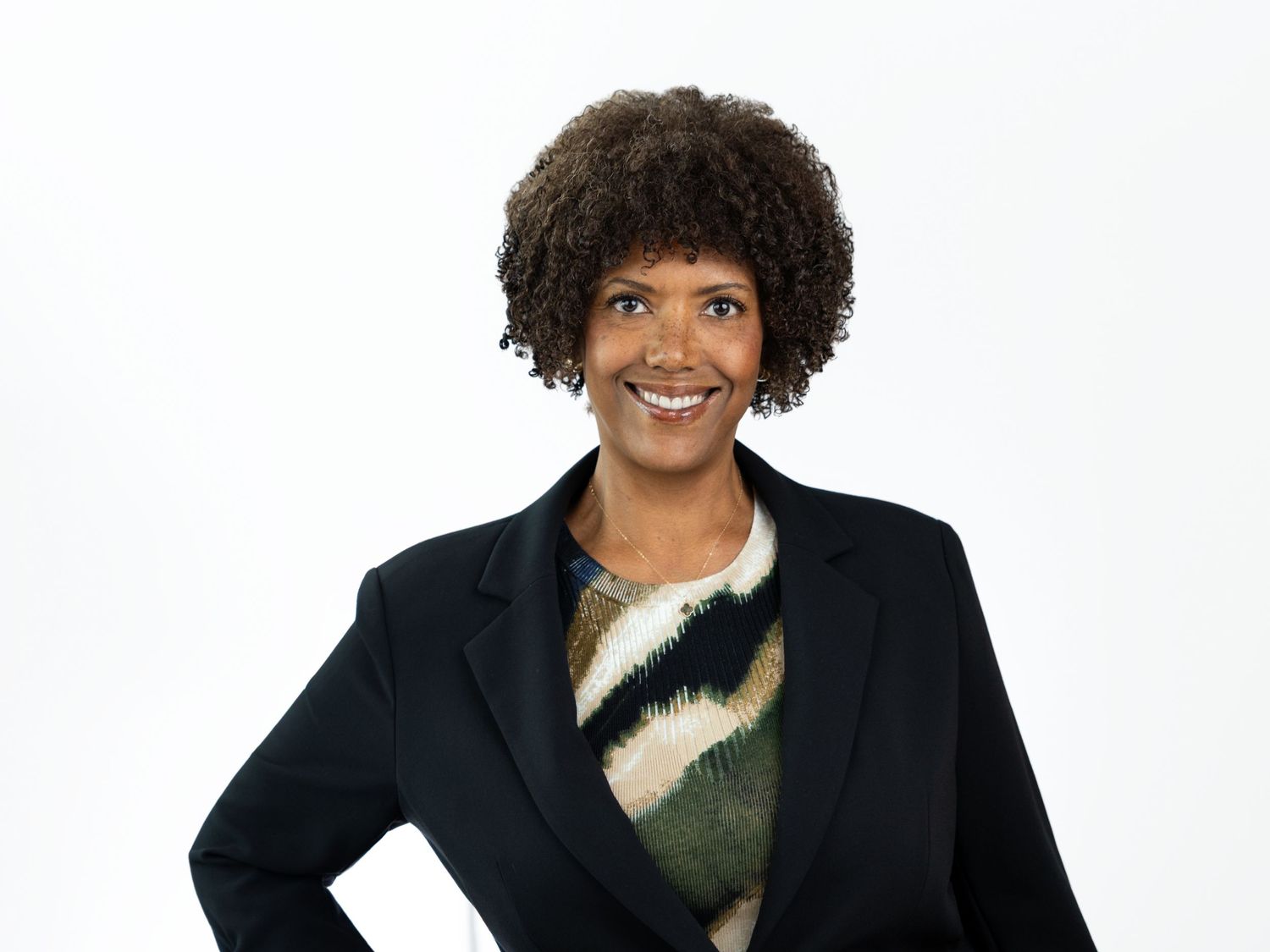 Smiling woman with short curly hair wearing a black blazer over a colorful patterned top against a white background.