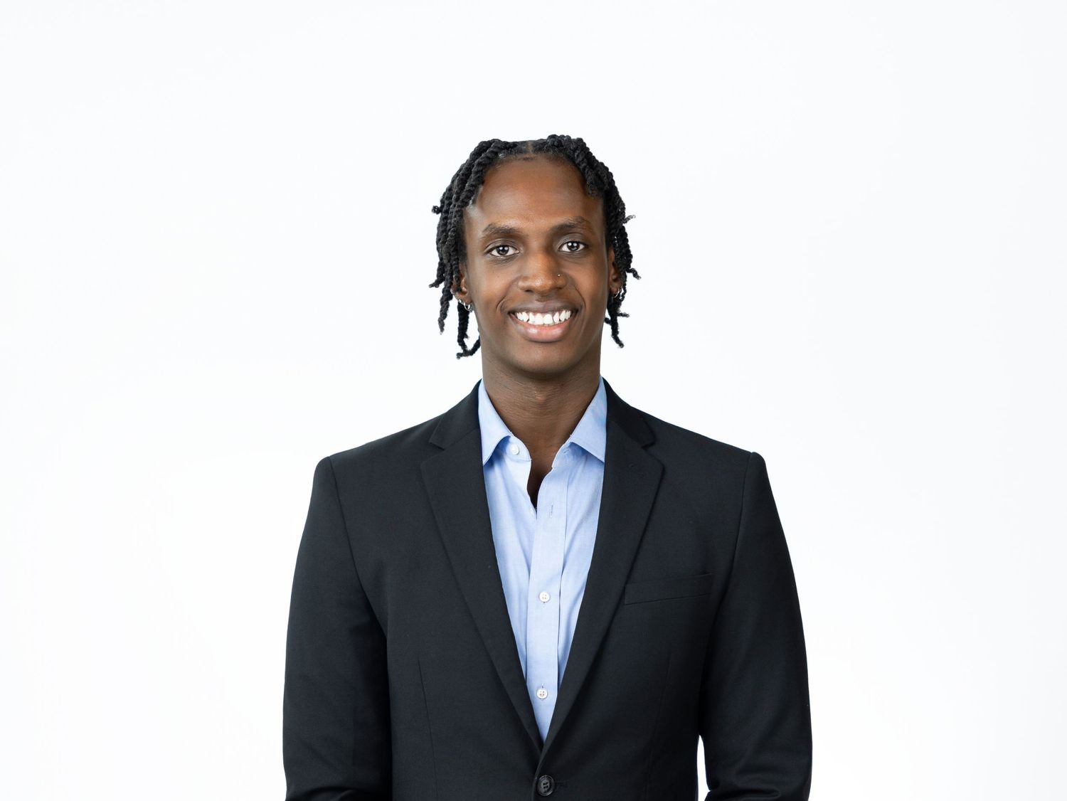 Smiling man with short twisted hair wearing a dark textured blazer over a light blue collared shirt against a white background.