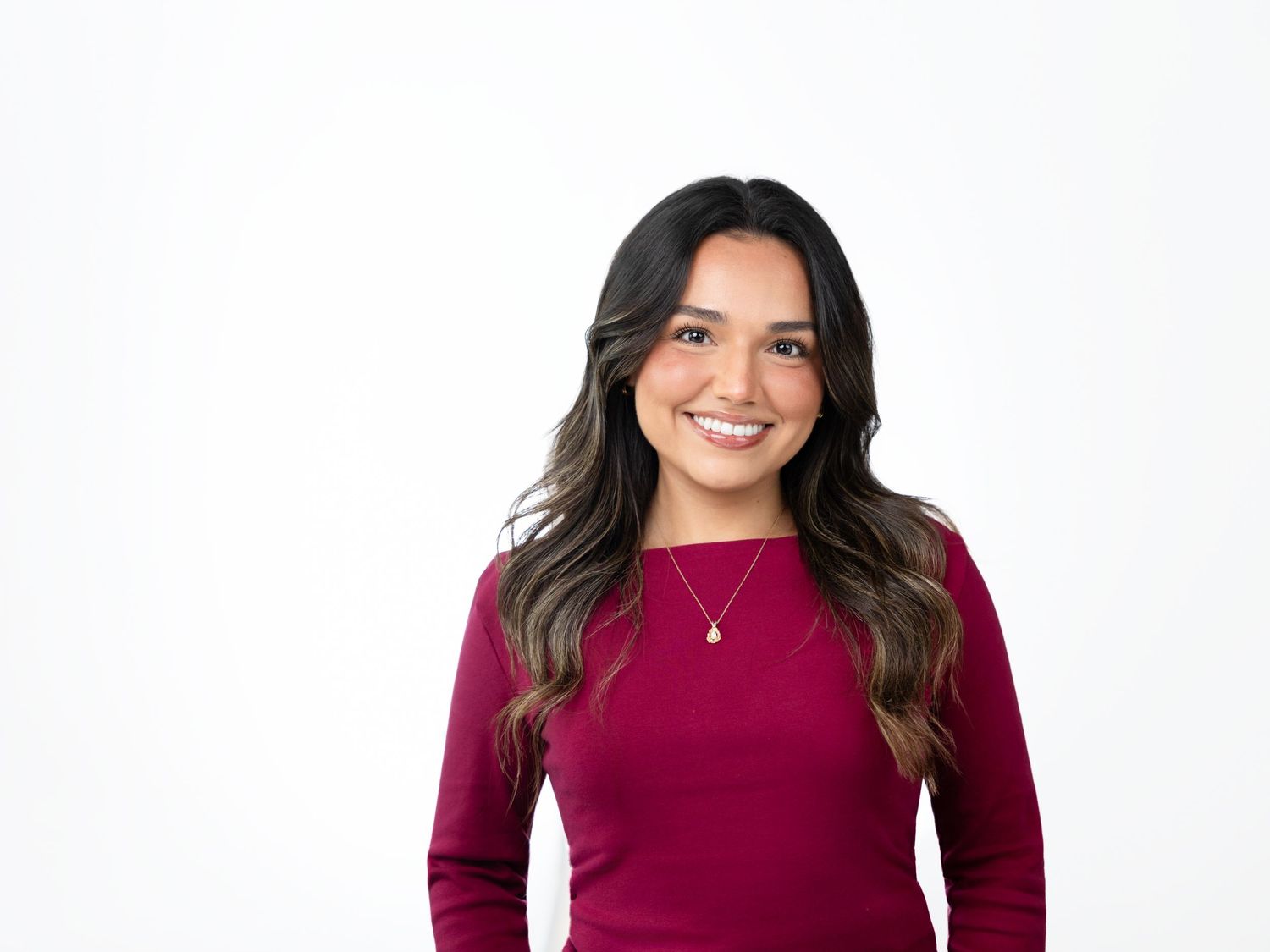 Smiling woman with long wavy dark hair wearing a burgundy long-sleeve top and a delicate pendant necklace against a white background.