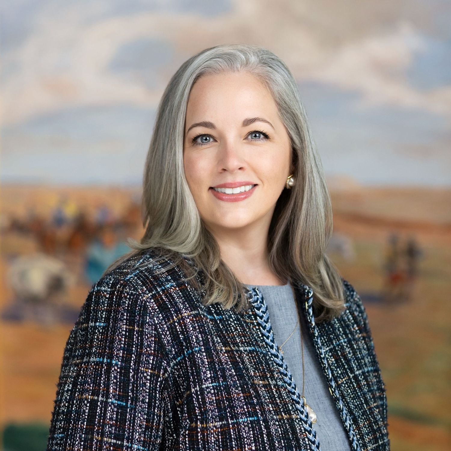 Smiling woman with long gray hair wearing a tweed jacket posing in front of a painted western landscape backdrop.