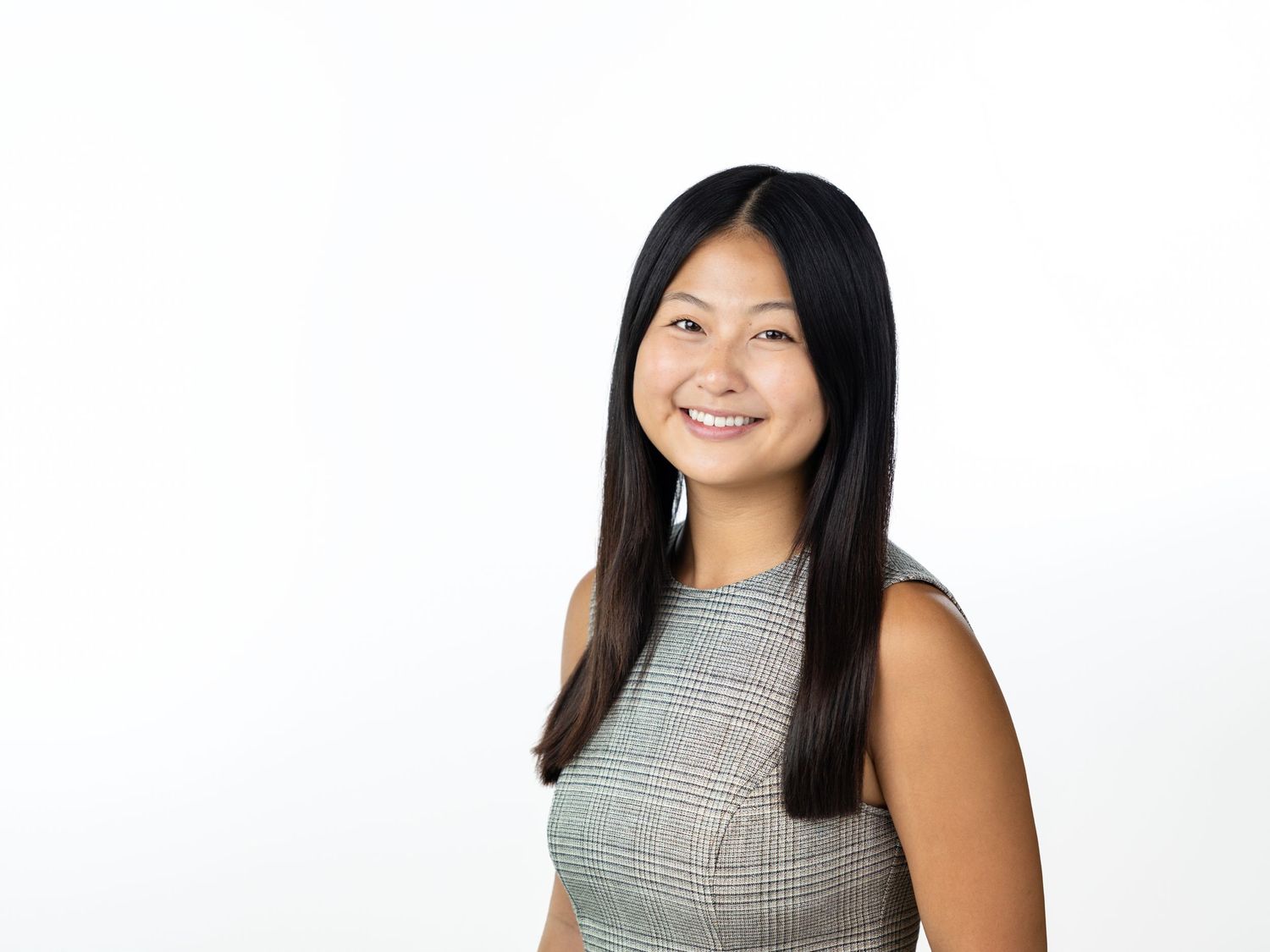 Smiling woman with long straight dark hair wearing a gray plaid sleeveless top against a bright white background.