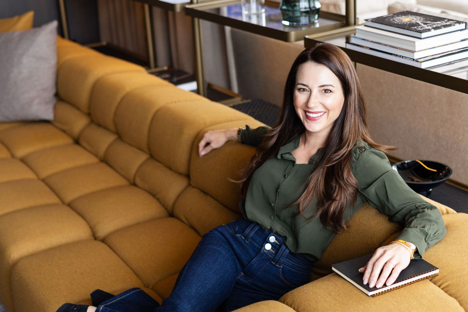 Smiling woman with long brown hair in a green blouse and jeans relaxes on a tufted mustard-yellow sofa holding a tablet.
