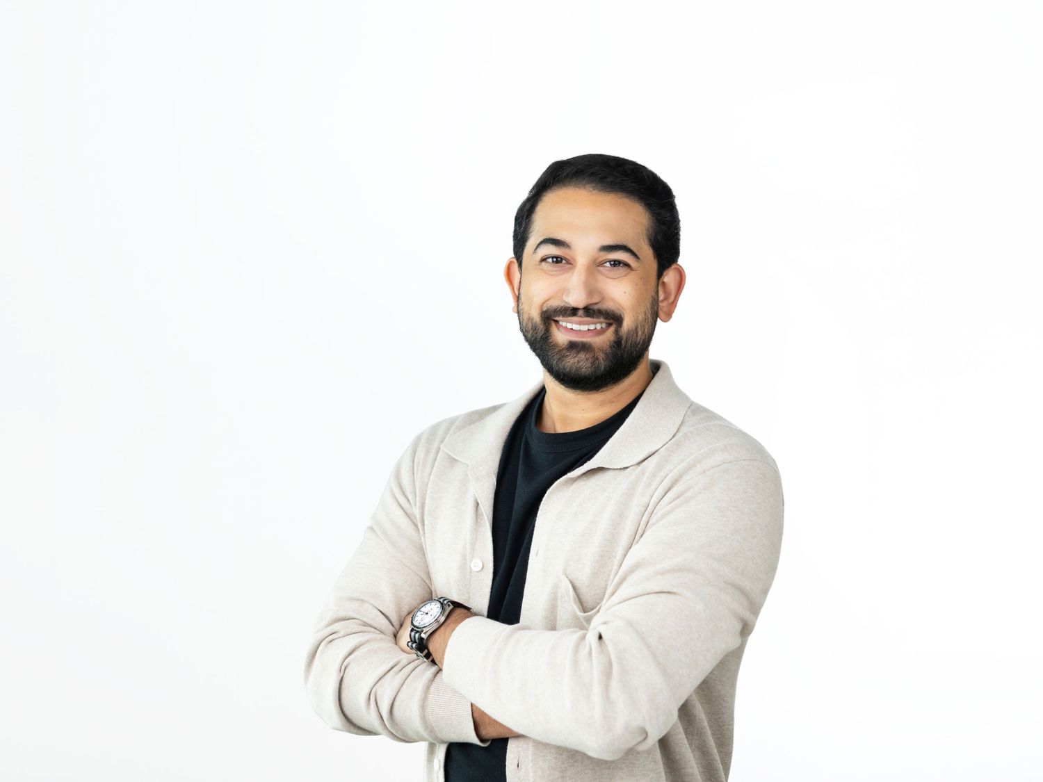 Bearded man with a friendly smile and crossed arms wearing a beige jacket over a black shirt and a wristwatch.