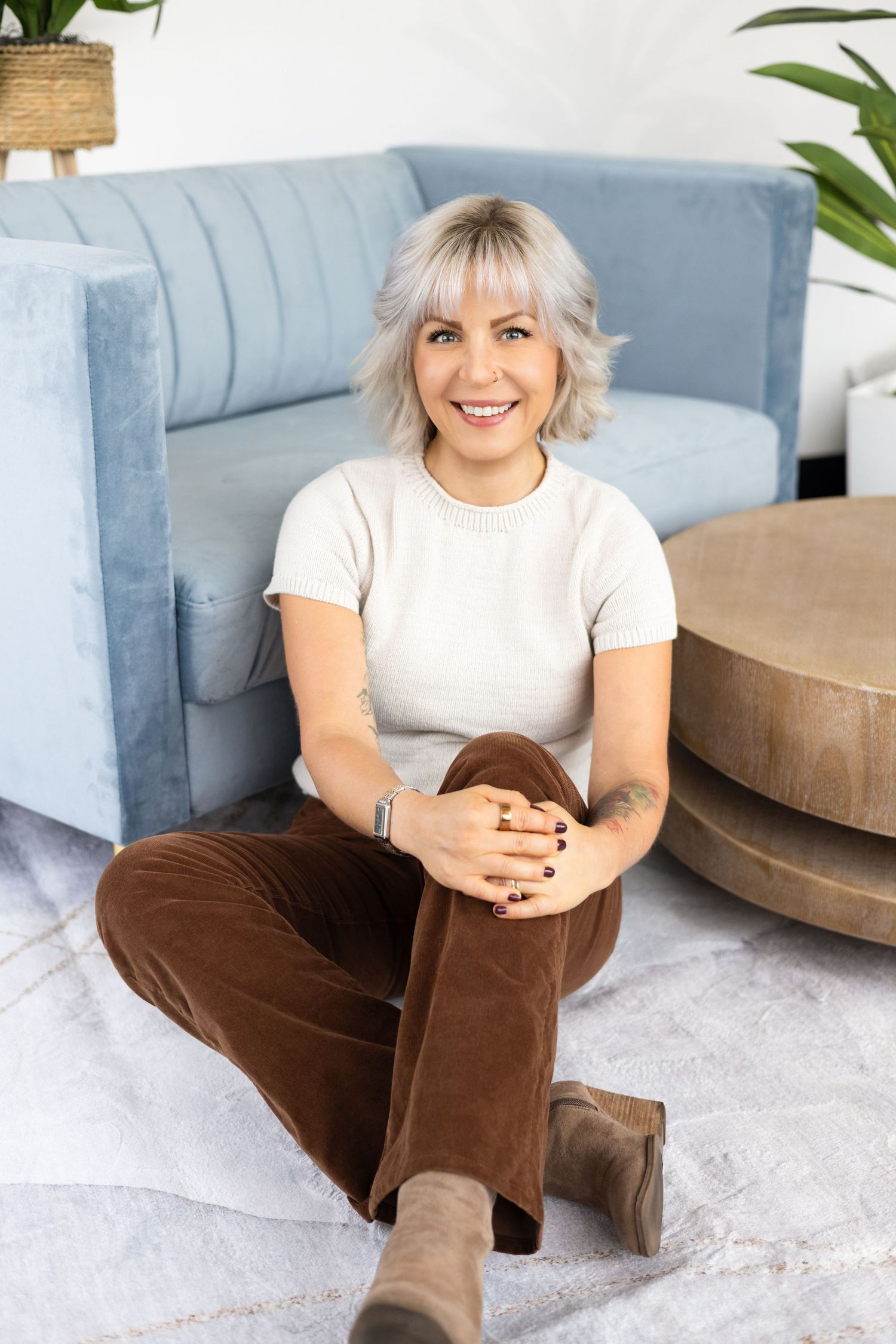 Smiling woman with short platinum hair sits on a rug in brown corduroy pants beside a blue sofa and wooden table.