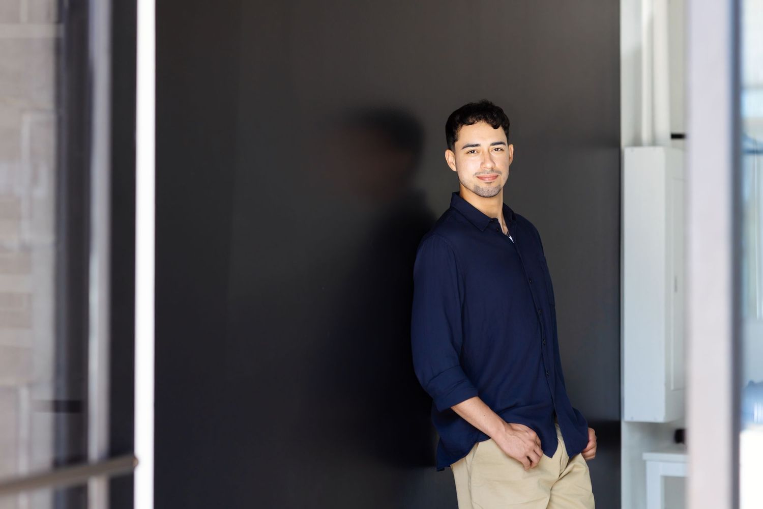 Man in a navy button-up shirt and khaki pants leans against a dark wall in a modern building with large windows.