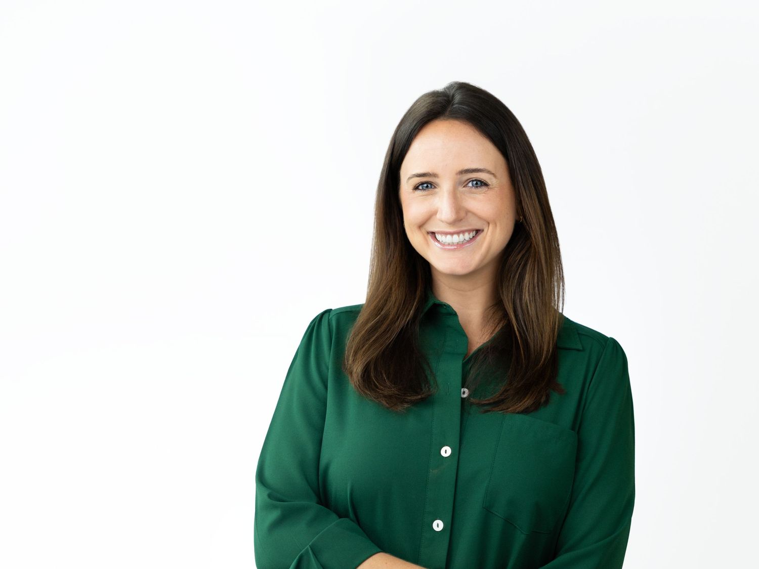 Smiling woman with long straight brown hair wearing a green button-up blouse standing against a bright white background.