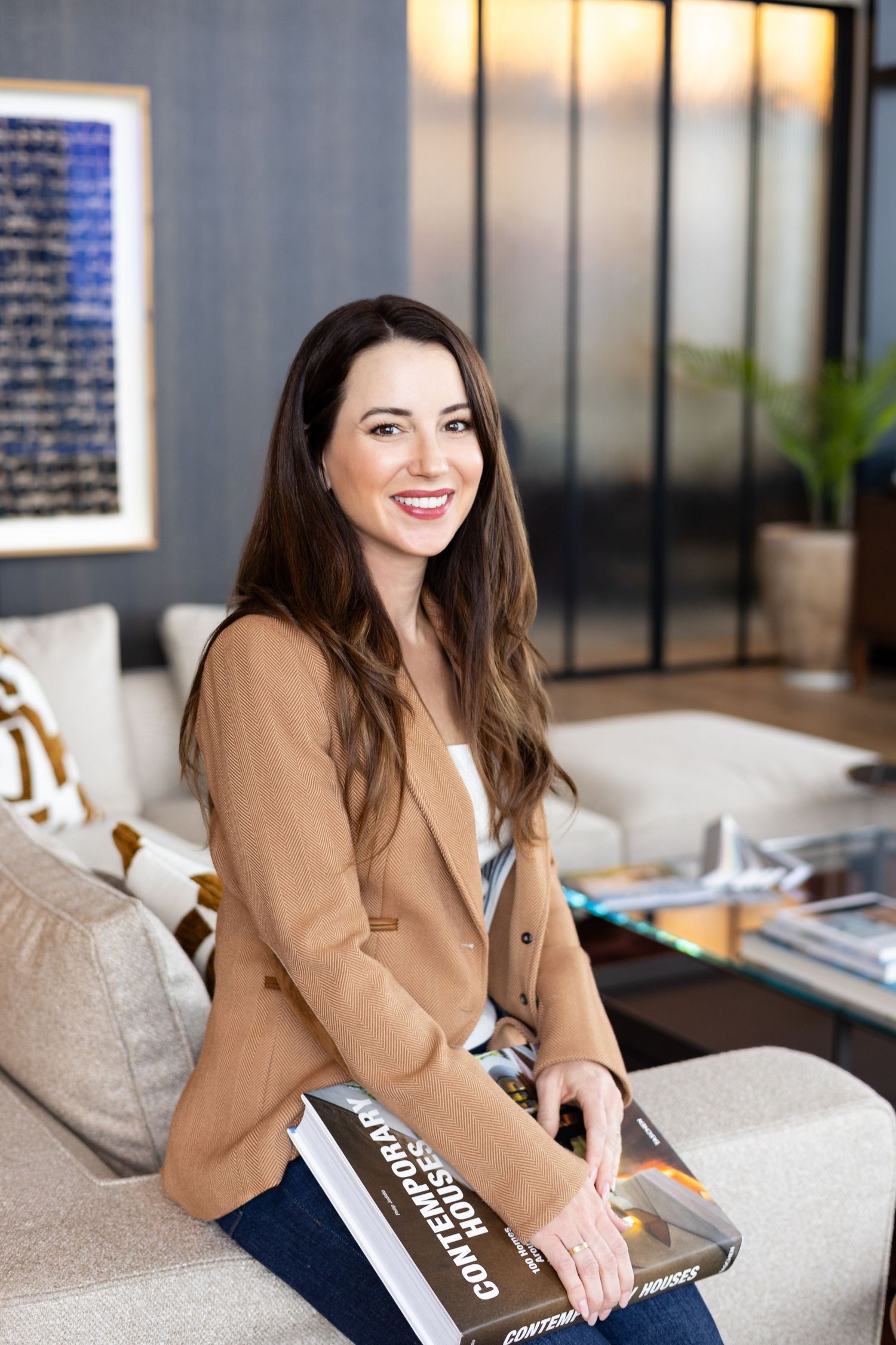 A smiling woman in a camel blazer sits on a sofa holding a large design book in a contemporary living room with framed art.