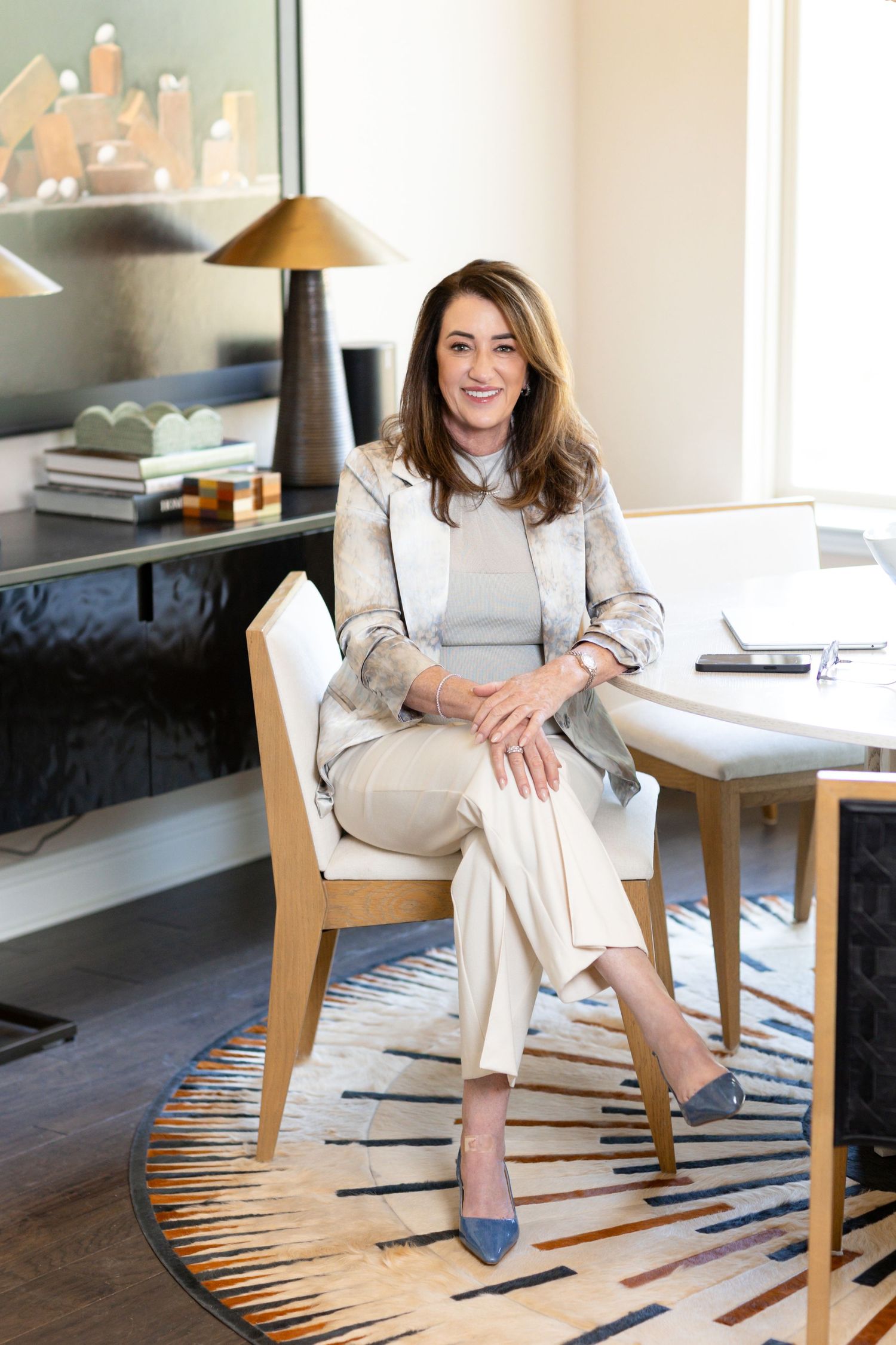 Confident woman in a blazer and wide-leg trousers sits on a wooden chair in a stylish office with a geometric rug and gold lamp.