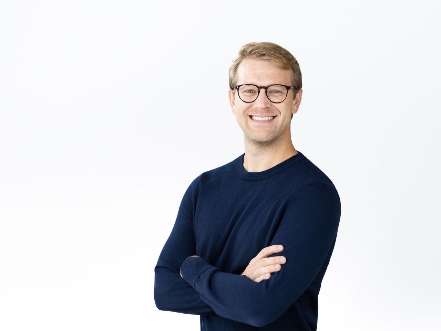 Smiling man with glasses and short blond hair stands with arms crossed in a navy blue sweater against a clean white background.