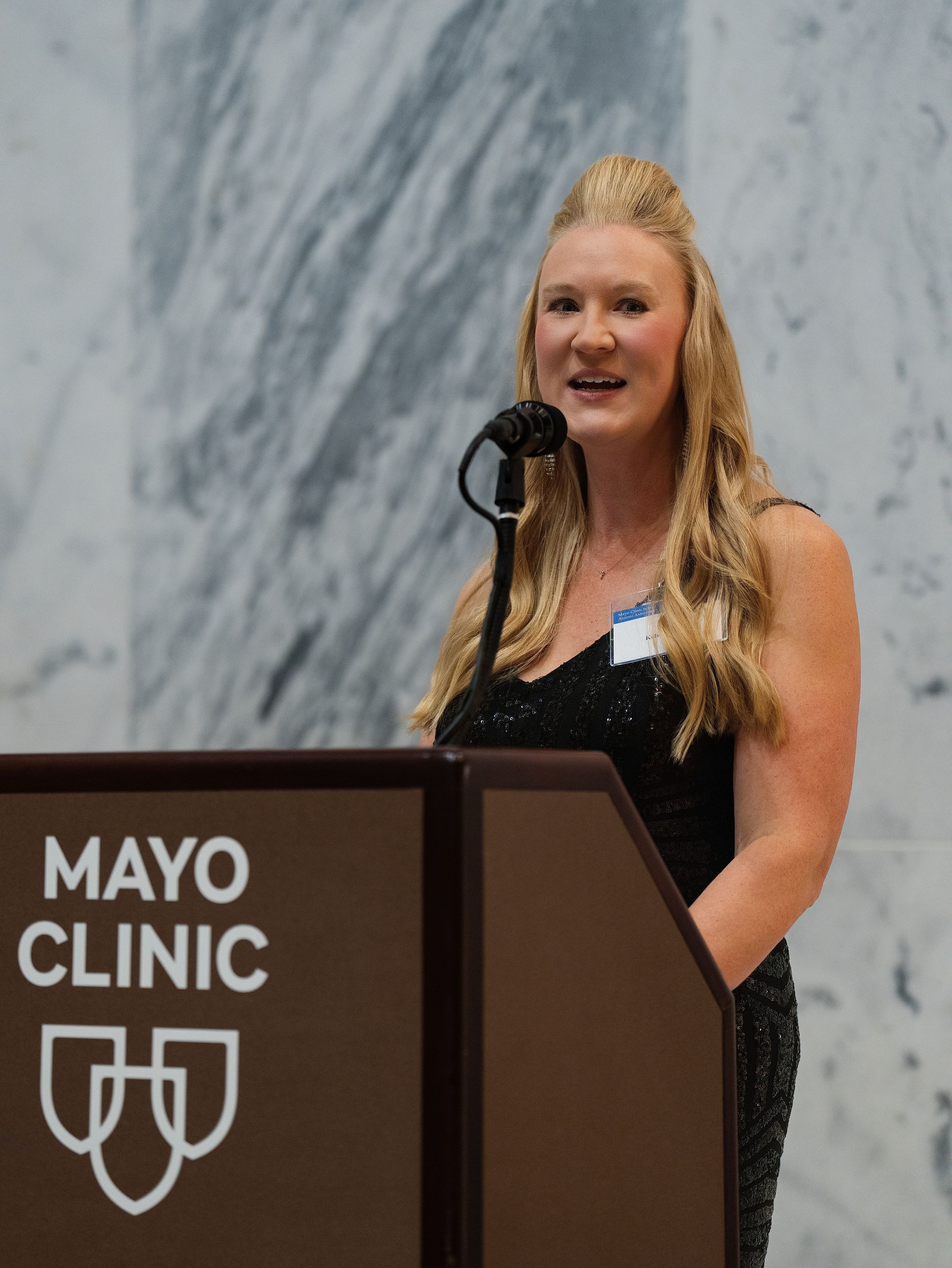 A woman speaking at a podium in front of a marble wall at a corporate event