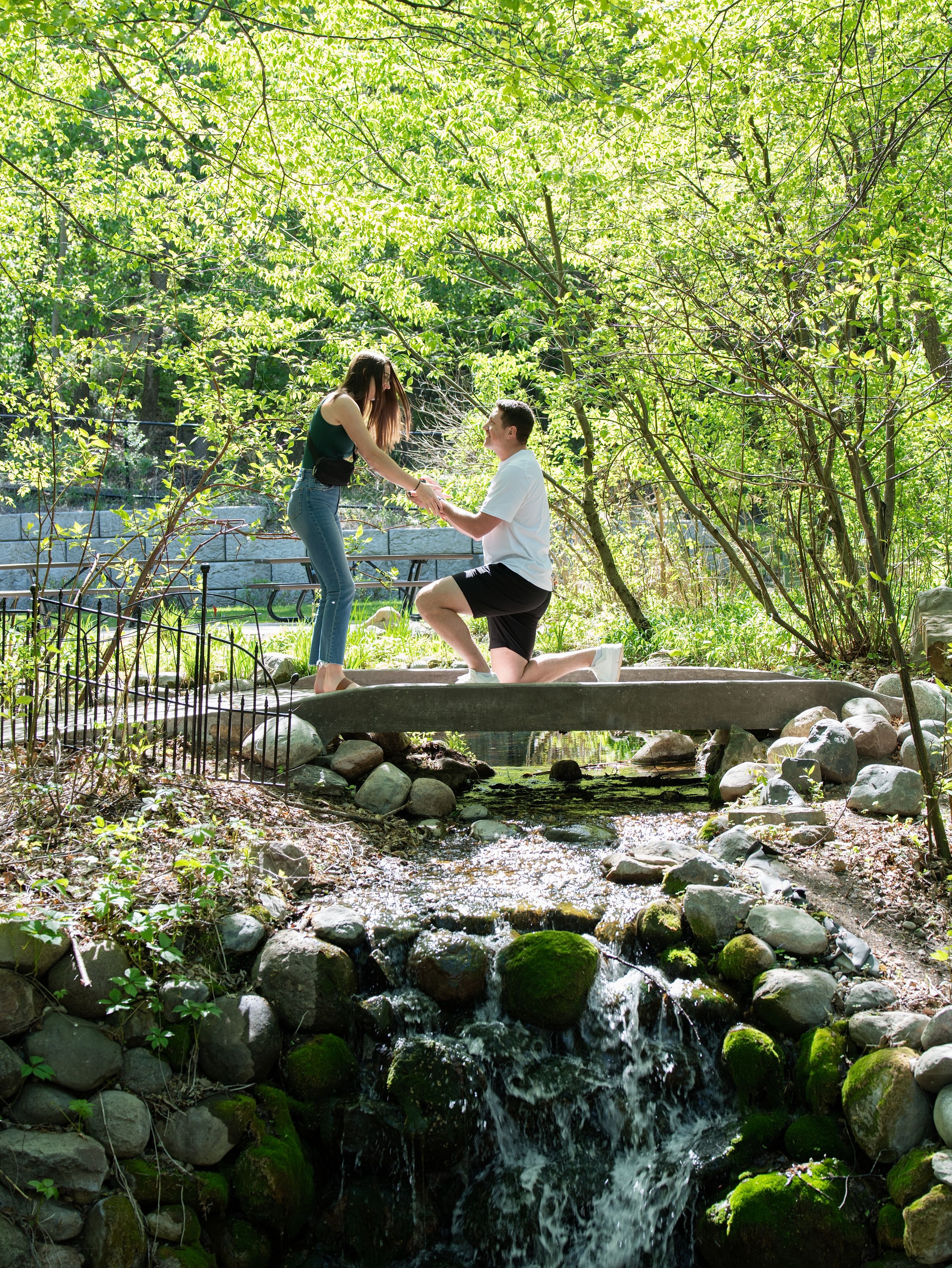 A man kneels to propose on a small stone footbridge above a mossy woodland stream