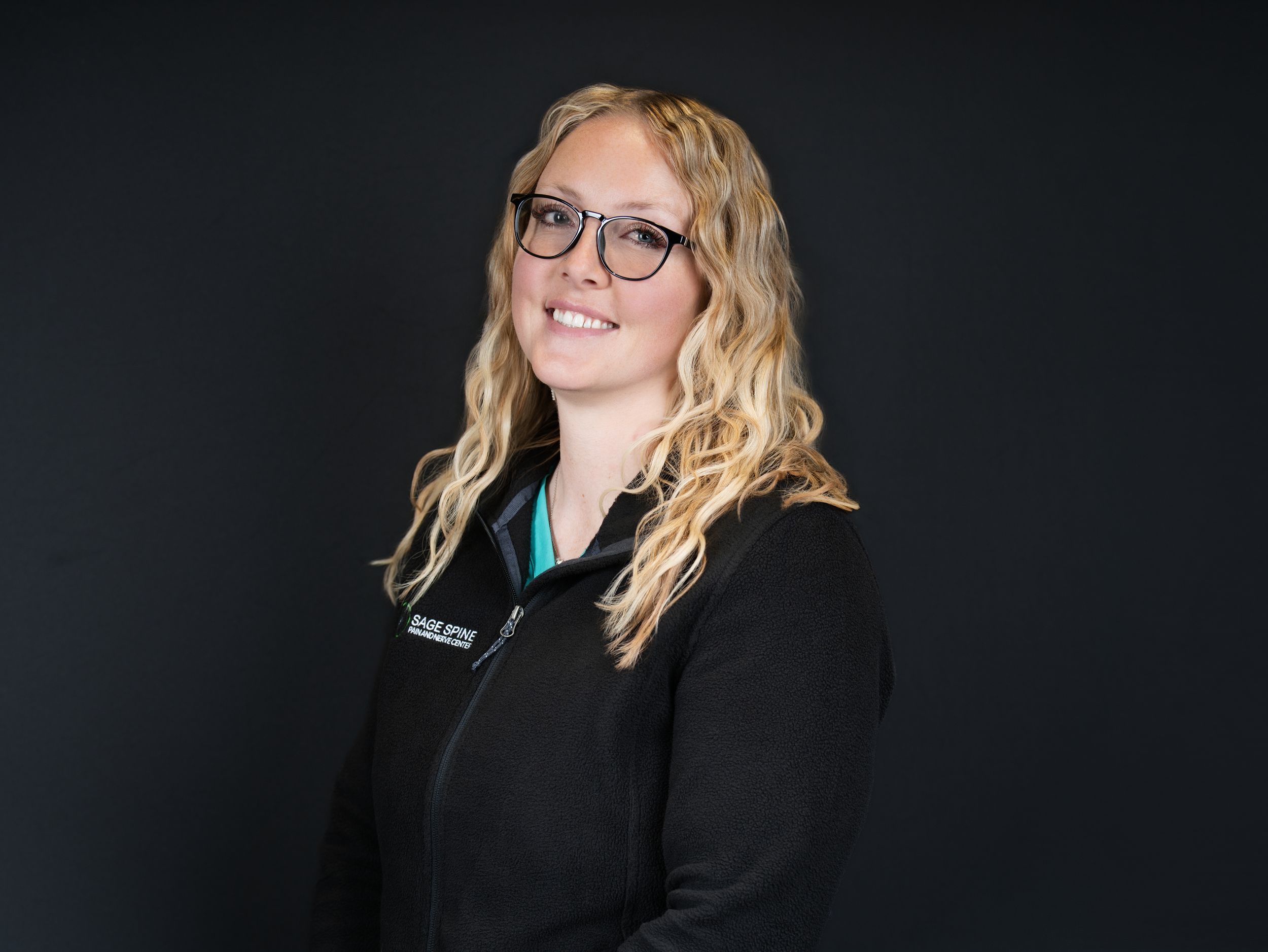 A smiling woman with curly blonde hair and glasses in a black zip-up jacket