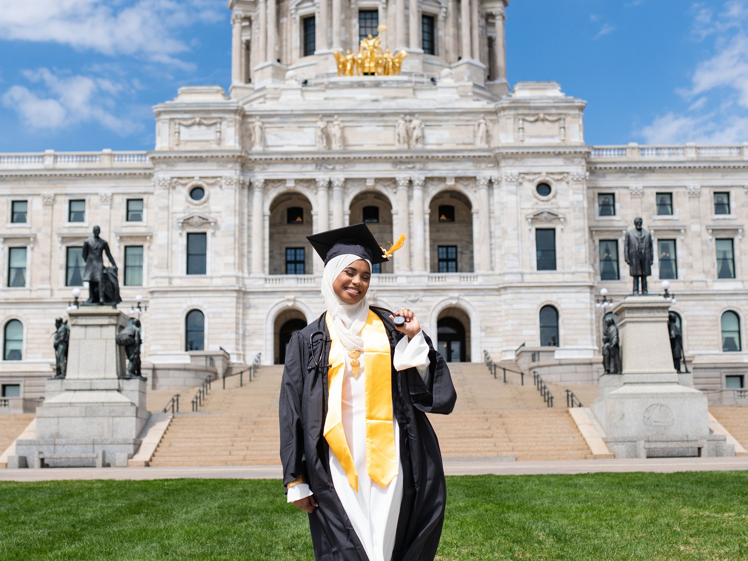 A graduate in a black gown and gold stole in front of the Minnesota State Capitol
