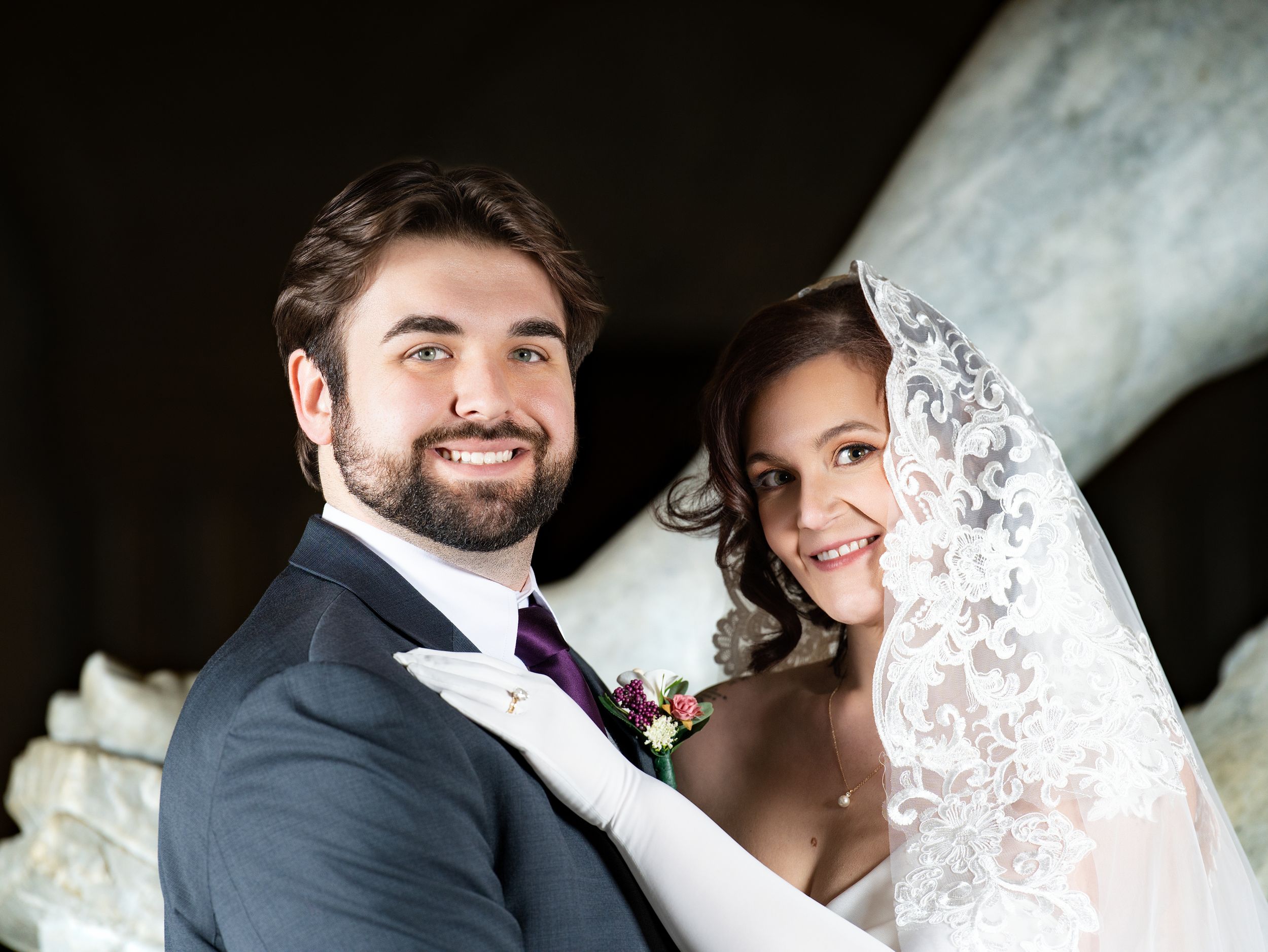 A groom and a bride in a lace veil and white gloves pose before a marble sculpture