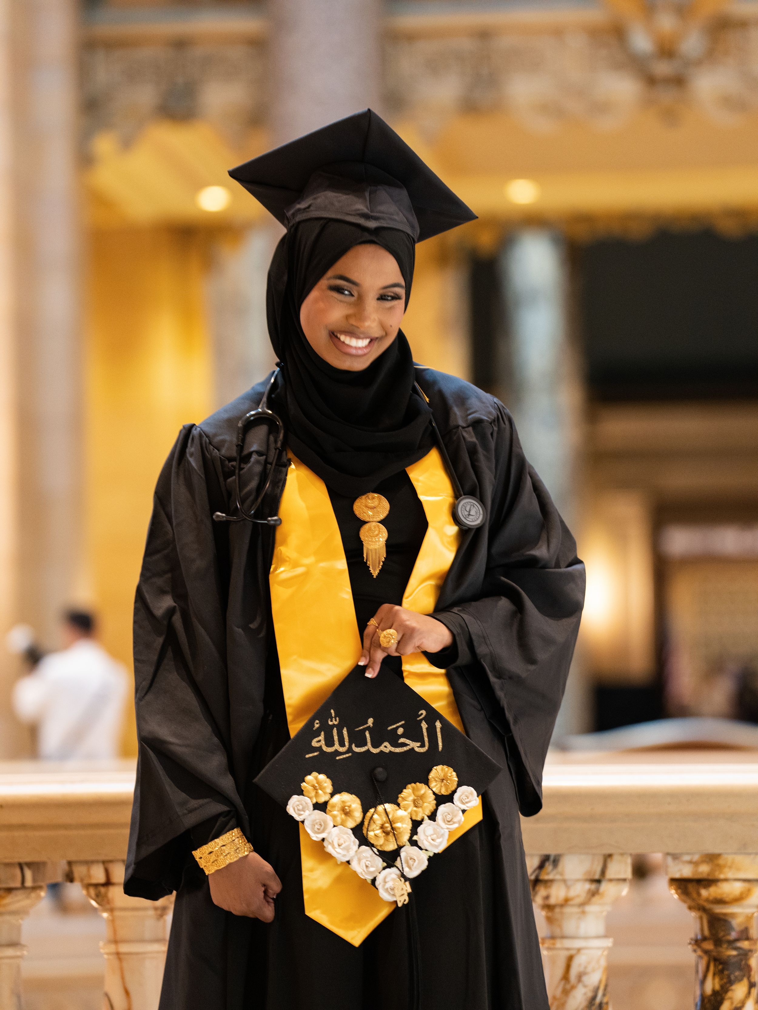 A graduate in a black gown and gold stole holding a decorated cap in an ornate hall
