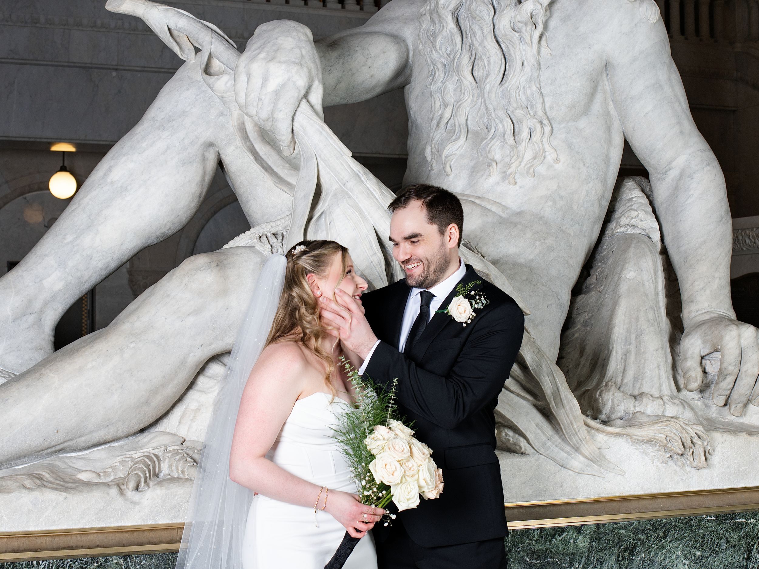A bride holding white roses touches the groom's face beside a large marble statue