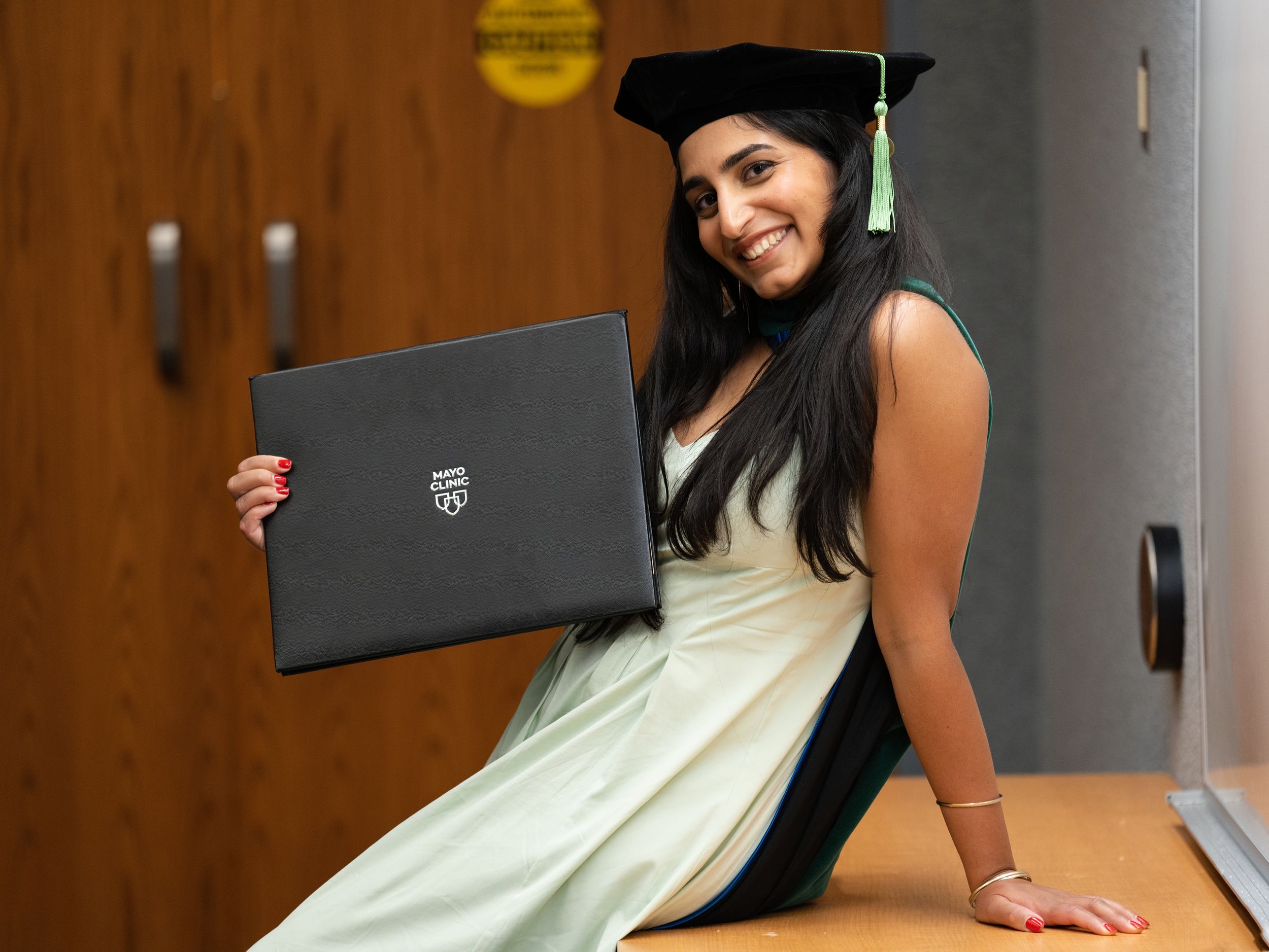A graduate in a doctoral cap sitting on a desk and holding up a diploma cover