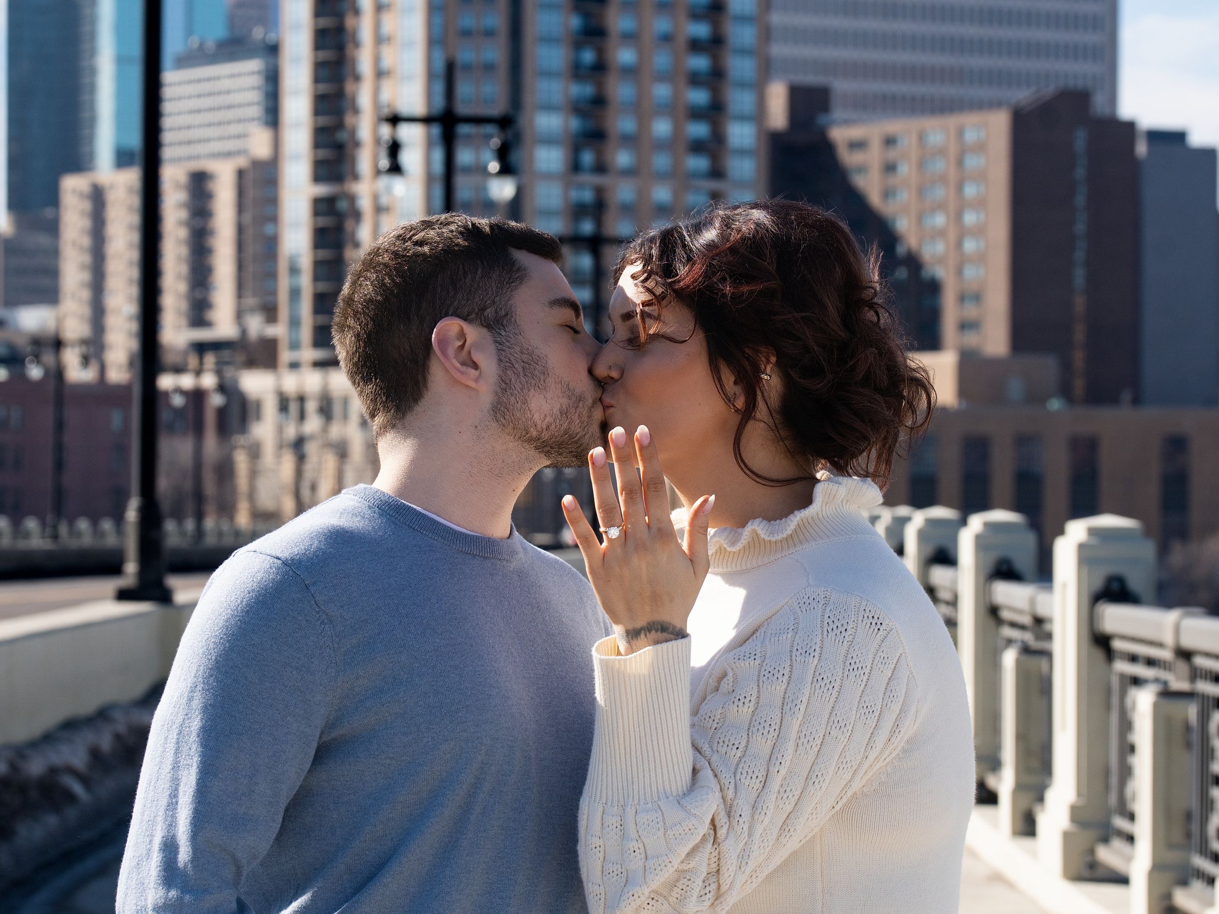 A newly engaged couple kisses as the woman shows her ring on a city bridge