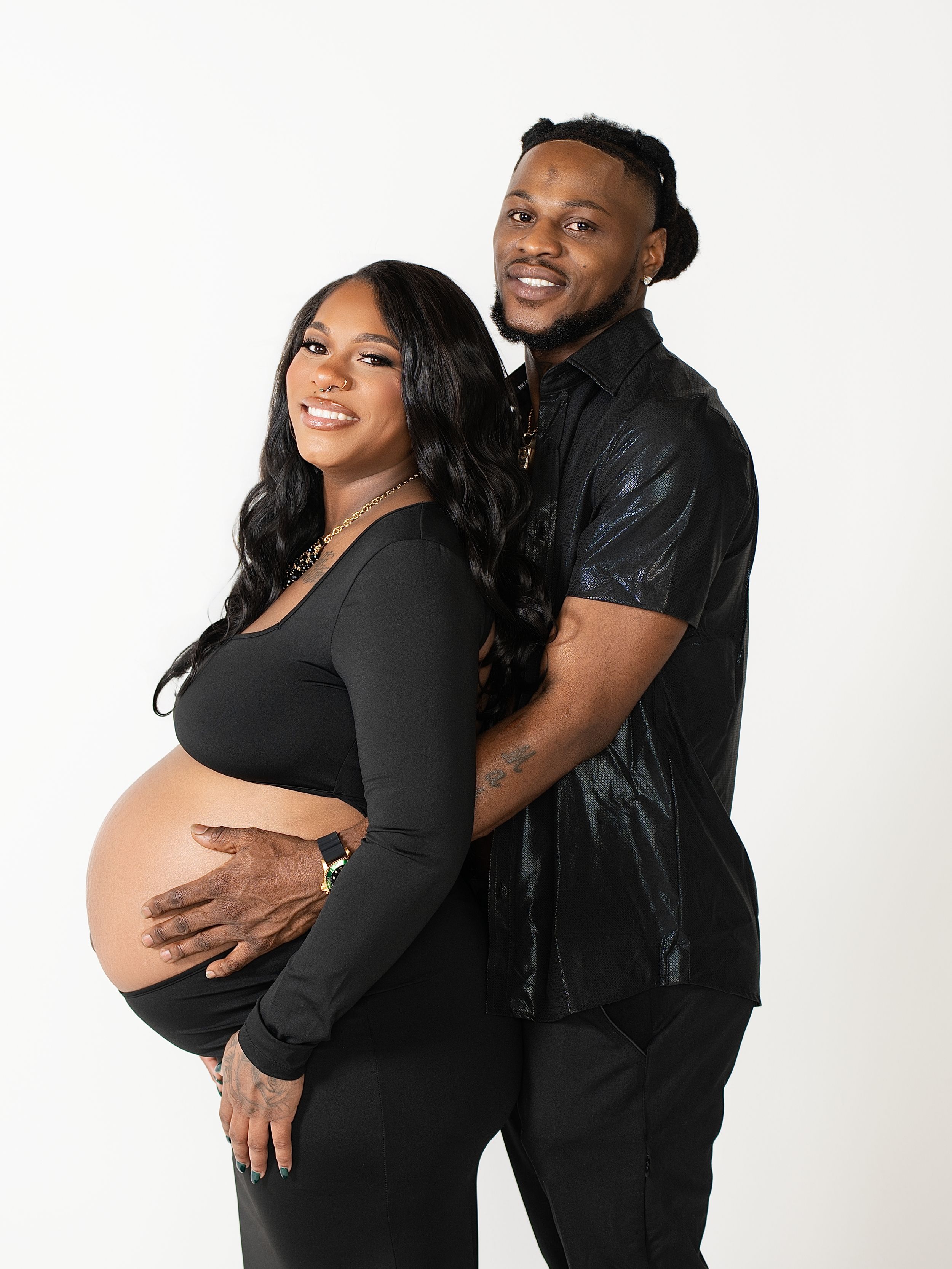 A man embracing his pregnant partner from behind in a bright white studio