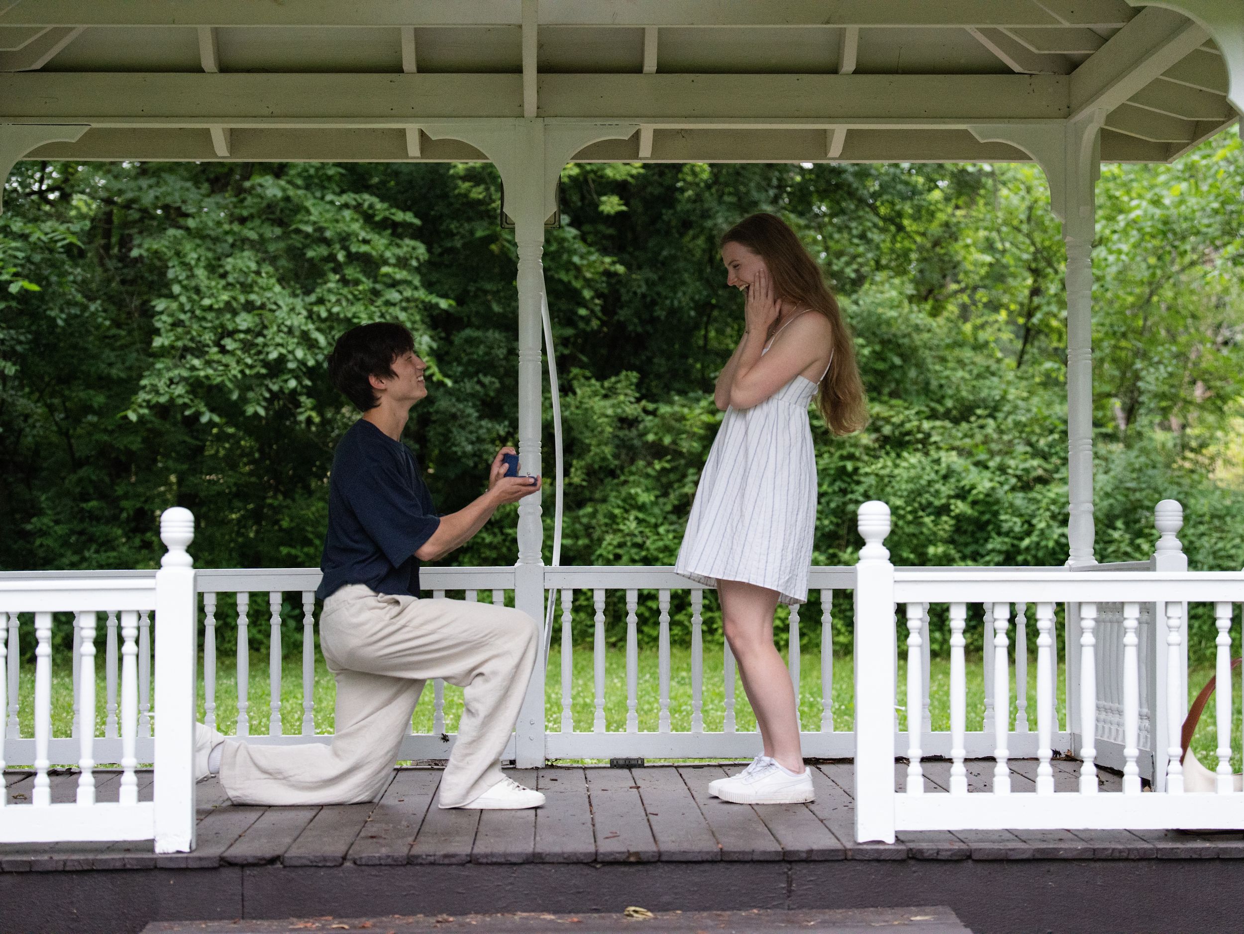 A man kneels to propose to a woman in a white sundress inside a white garden gazebo