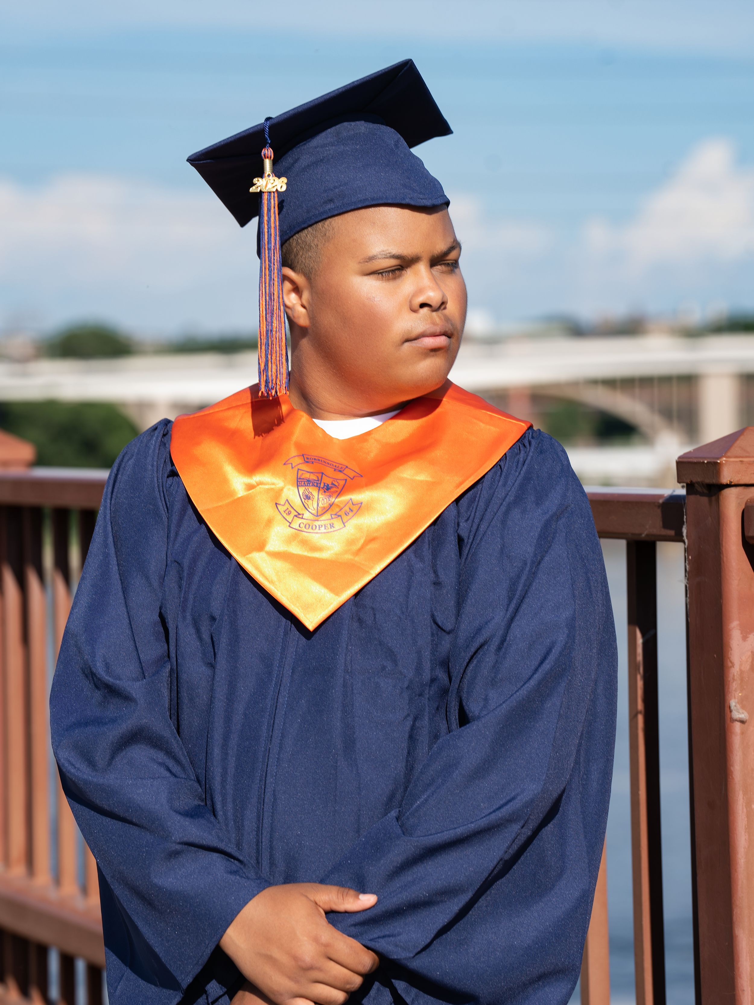A graduate in a navy gown and orange stole standing on a bridge above a river