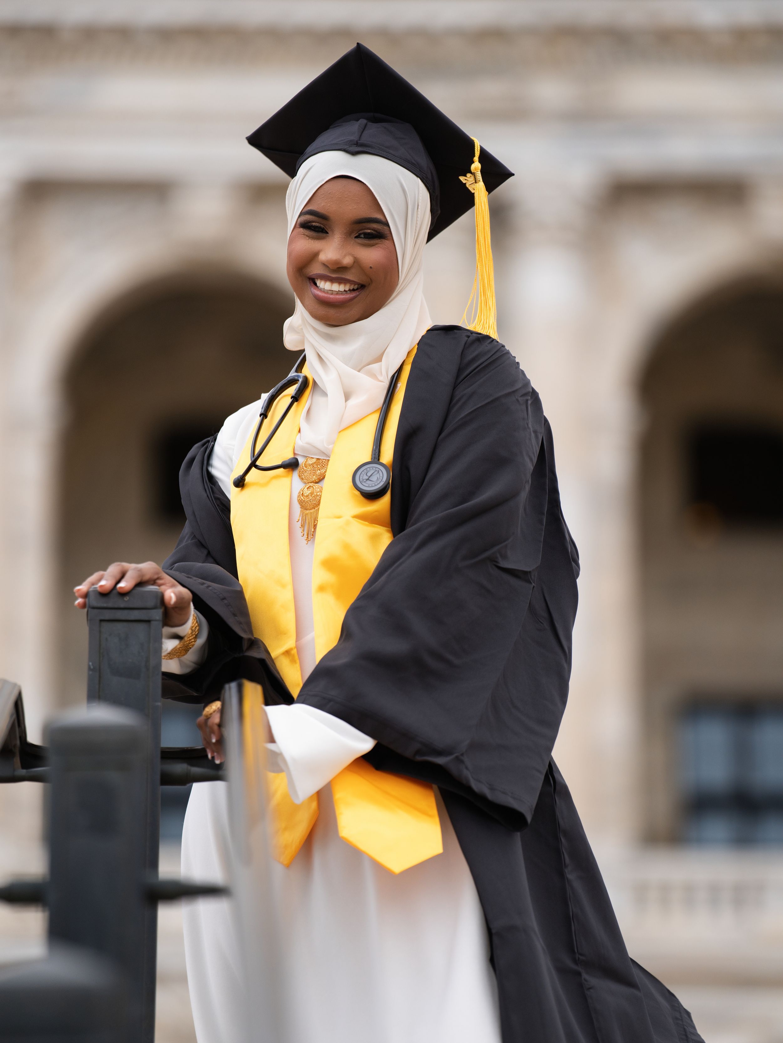 A graduate in a black gown and gold stole with a stethoscope smiling beside stone arches