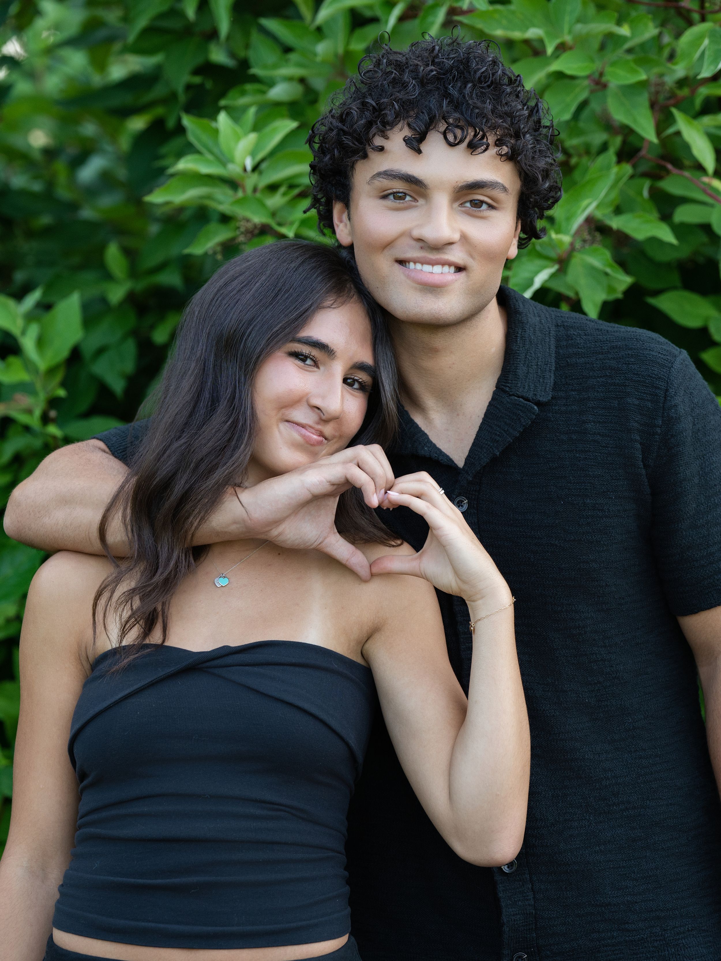 A young couple smiling as the woman makes a heart shape with her hands among green leaves