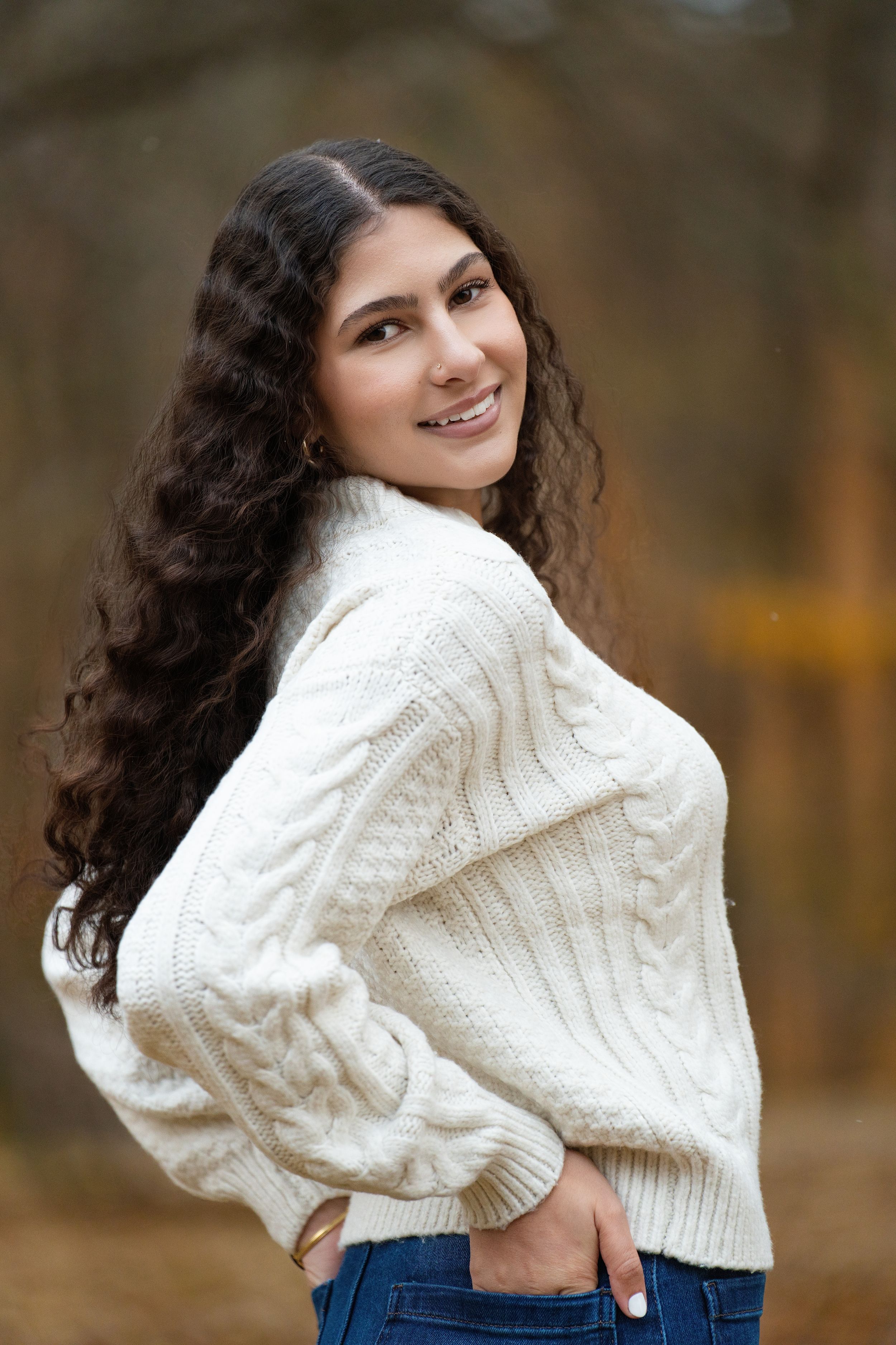 A young woman in a cream cable-knit sweater looking over her shoulder in autumn woods