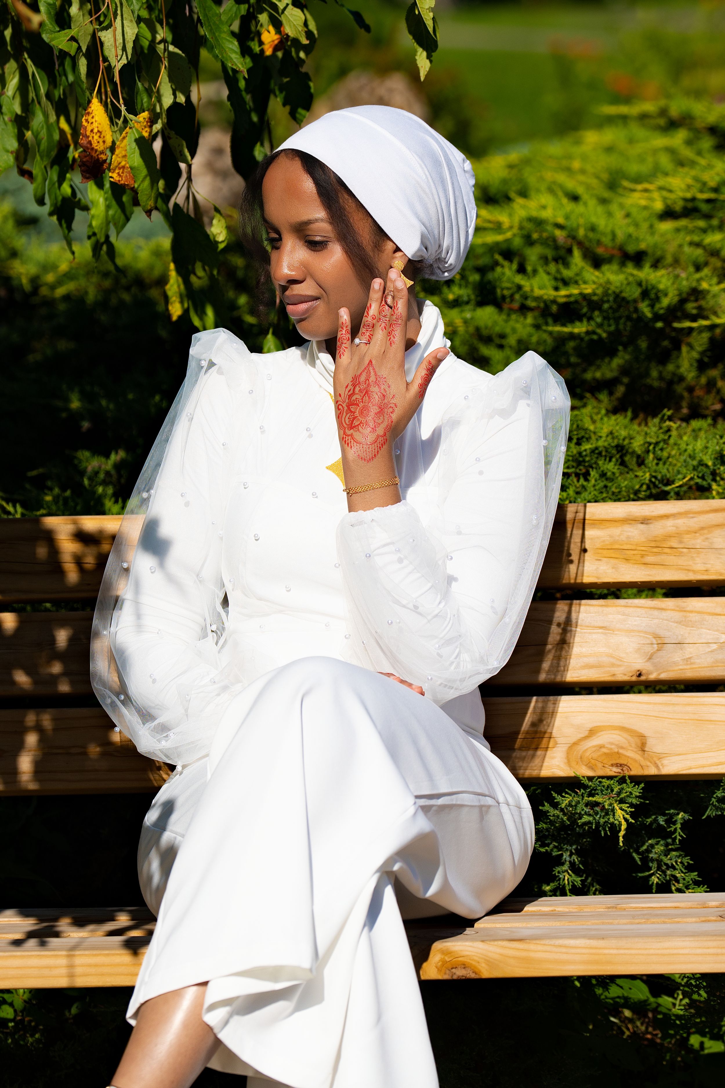 A woman in a white outfit and white head wrap sitting on a wooden bench among greenery