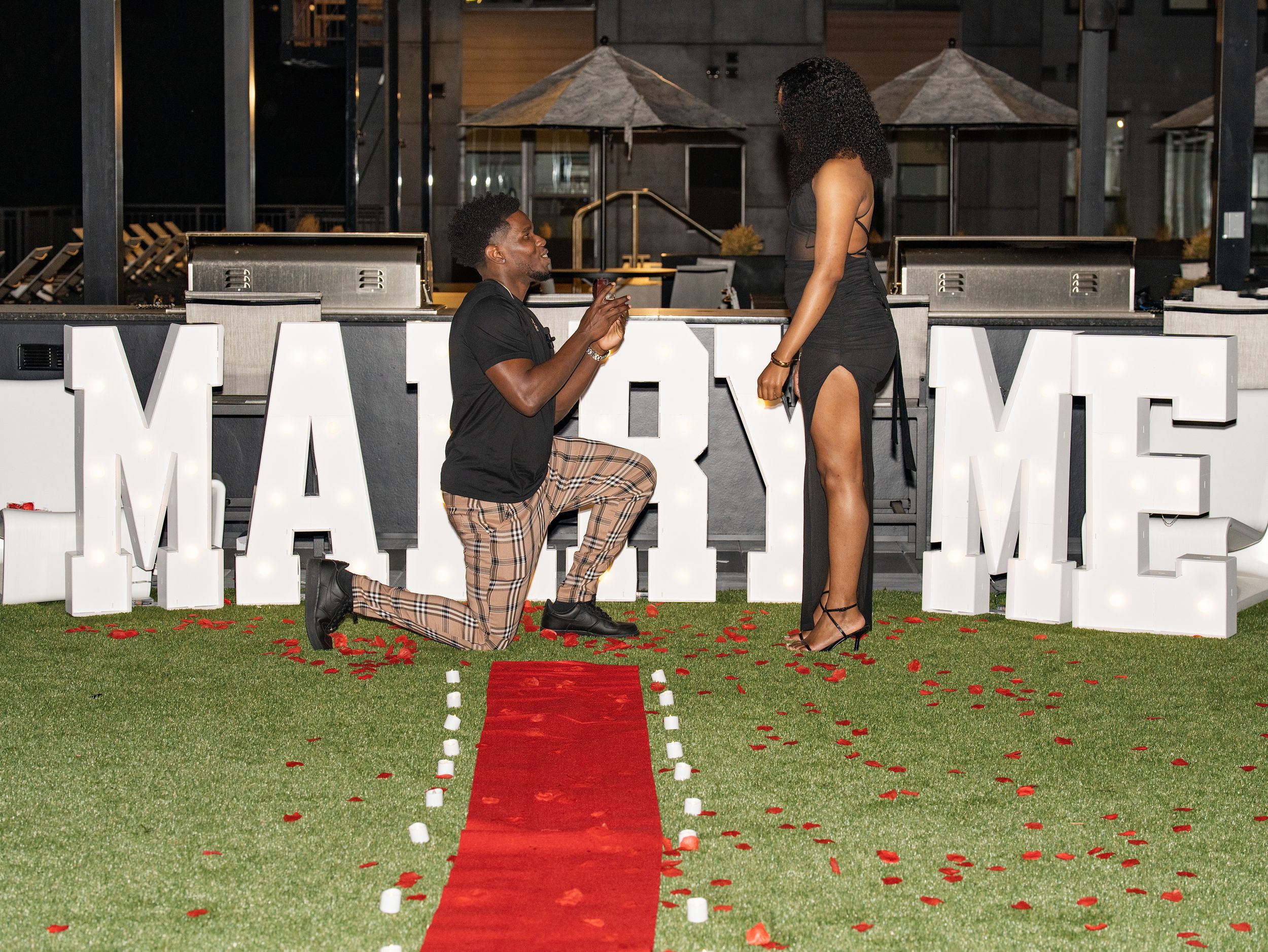 A man kneels to propose to a woman before lit MARRY ME letters and a red carpet