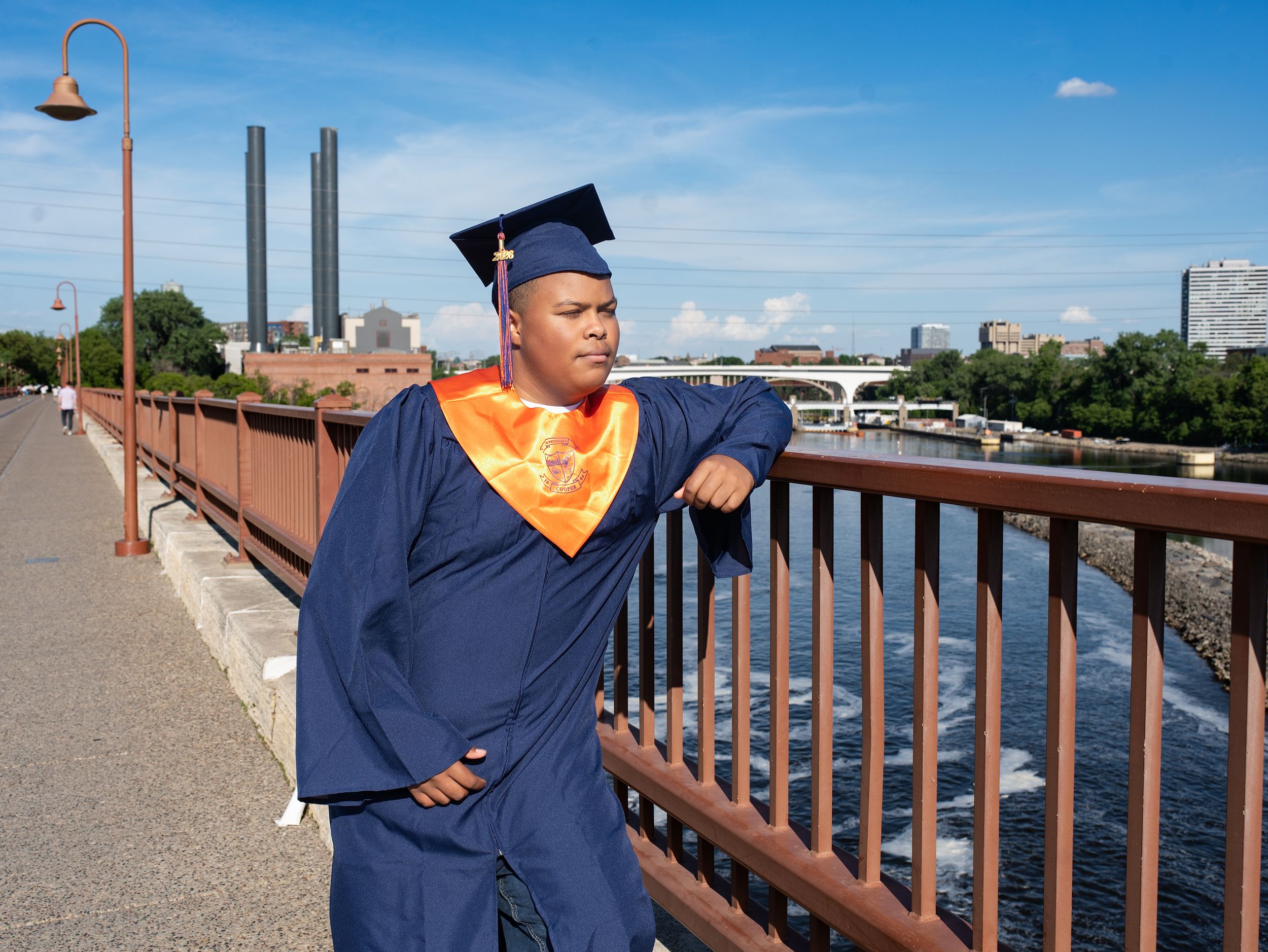 A graduate in a navy gown and orange stole leaning on a bridge railing above a river