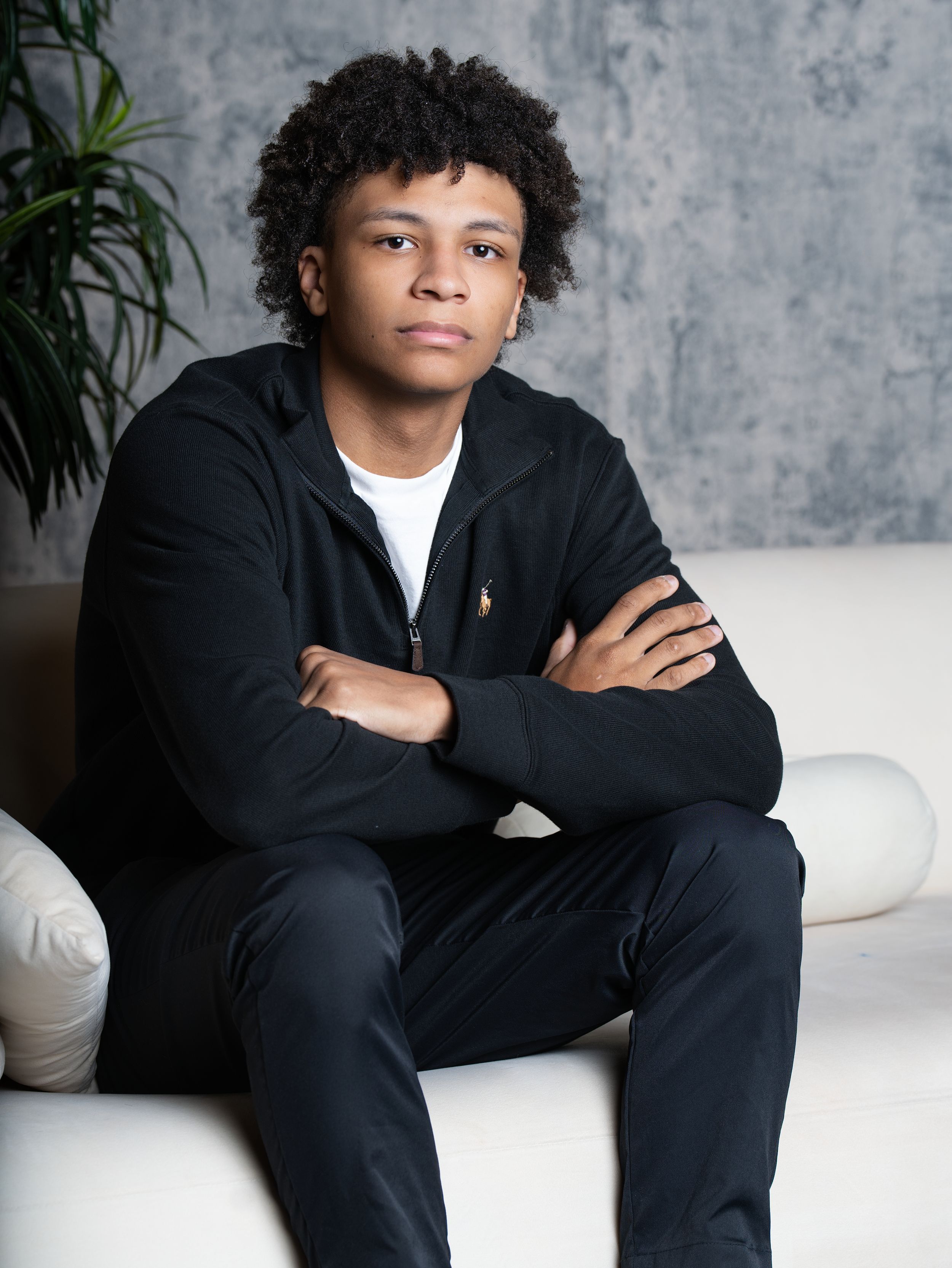 A young man in a black zip jacket sitting with arms crossed on a white studio sofa